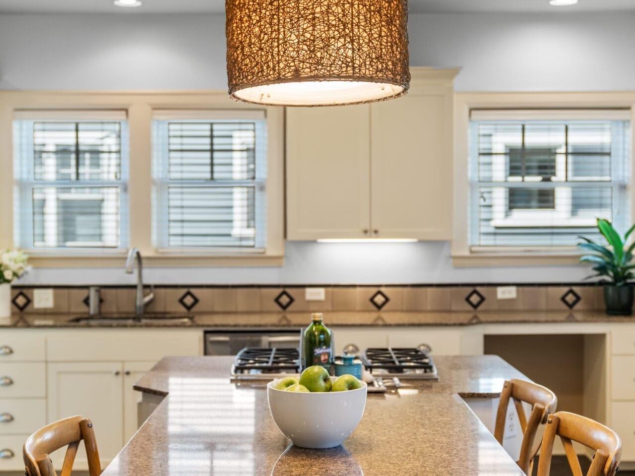 A modern kitchen with a large island countertop, wicker chairs, and a fruit bowl in the center. A woven pendant light hangs above, and the room is illuminated by natural light from windows in the background.