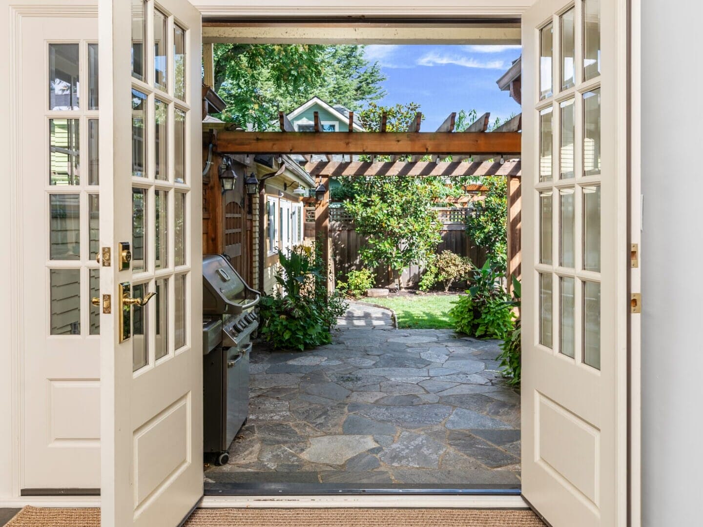 Open double doors lead to a stone patio with a grill on the left. A pergola with hanging plants and greenery is in the yard, with a vibrant green lawn and trees under a clear blue sky in the background.