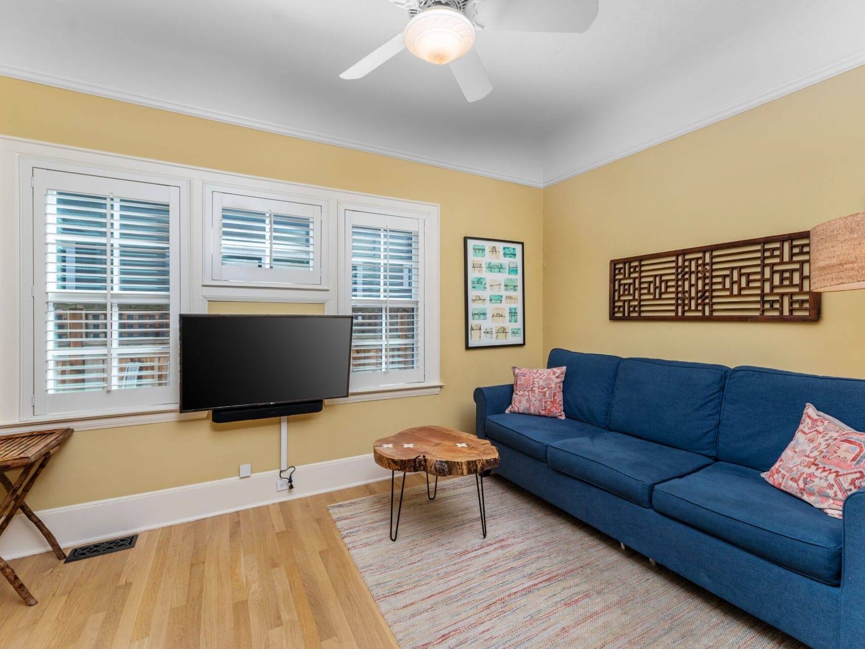 A cozy living room with a blue sofa, adorned with pink and white patterned pillows. A wooden coffee table is in front of the sofa, and a TV is mounted on the wall. The room features yellow walls, hardwood floors, and a ceiling fan.