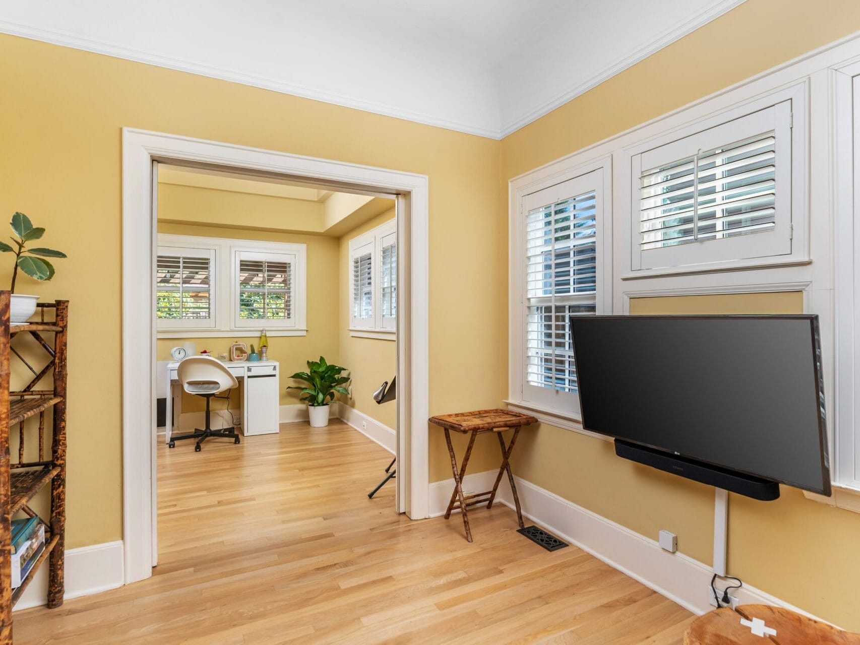 A cozy, light-filled room with yellow walls and hardwood floors. It features a wall-mounted TV, a wooden bookshelf with a plant, and a view into a small office area with a white desk and chair, set against large windows with blinds.