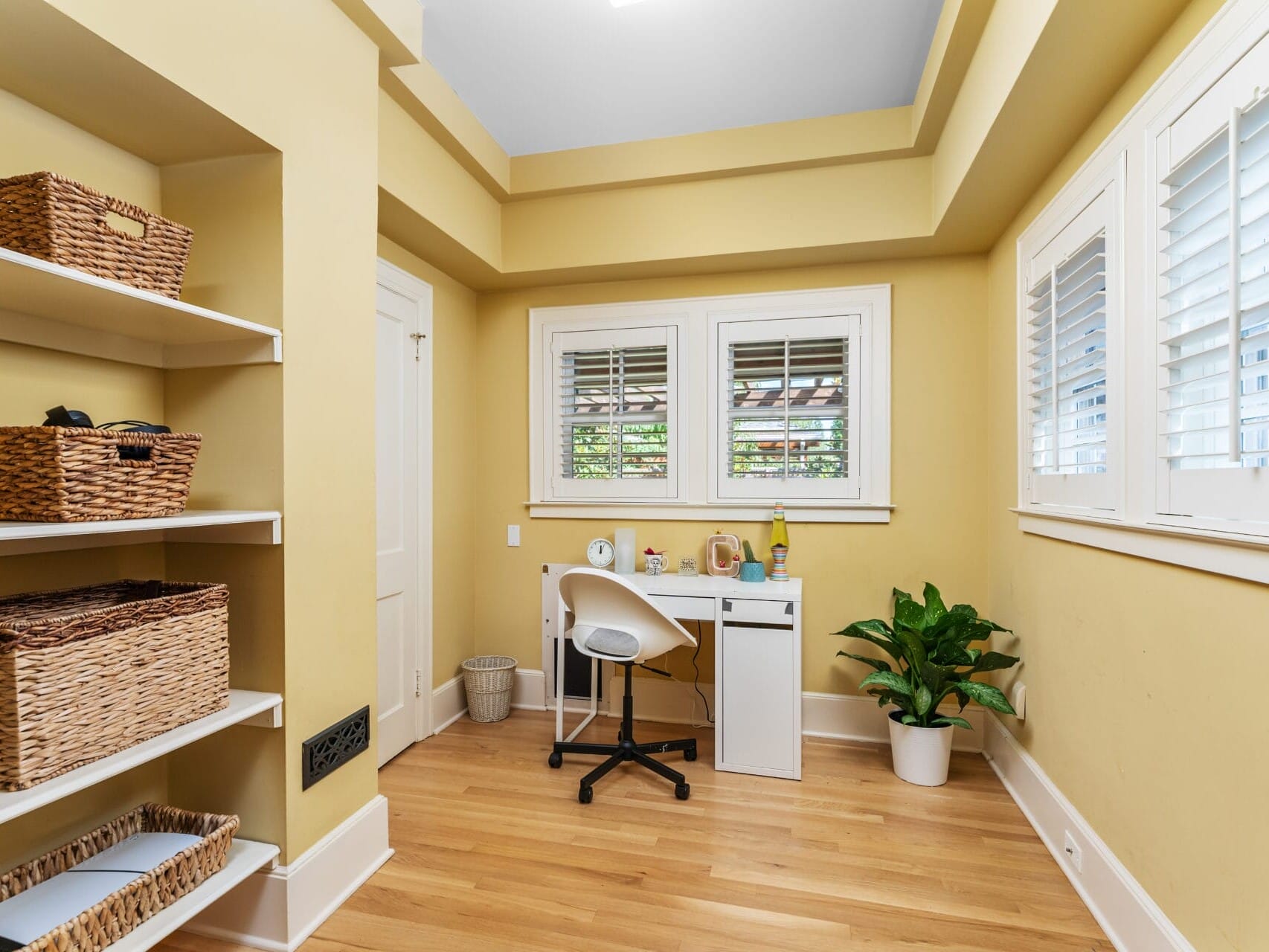 A small, neatly organized home office with yellow walls and wooden floors. It features a white desk with a white chair, a plant in the corner, and shelves with wicker baskets. Large windows with blinds let in natural light.