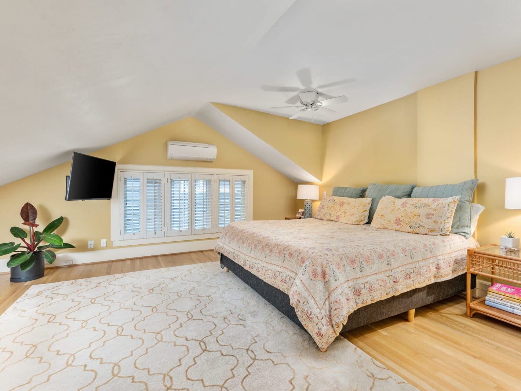 A cozy attic bedroom with sloped ceilings features a large bed with patterned bedding, two bedside lamps, and a potted plant. A wall-mounted TV is above a window with shutters. The room has wooden floors and a beige patterned rug.