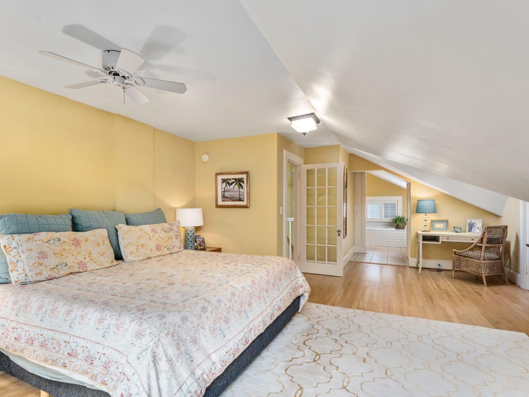 A cozy attic bedroom with a large bed adorned with floral bedding under a ceiling fan. Theres a beige rug on the wooden floor. In the corner is a desk with a chair near a window, separated by French doors, and soft yellow walls throughout.