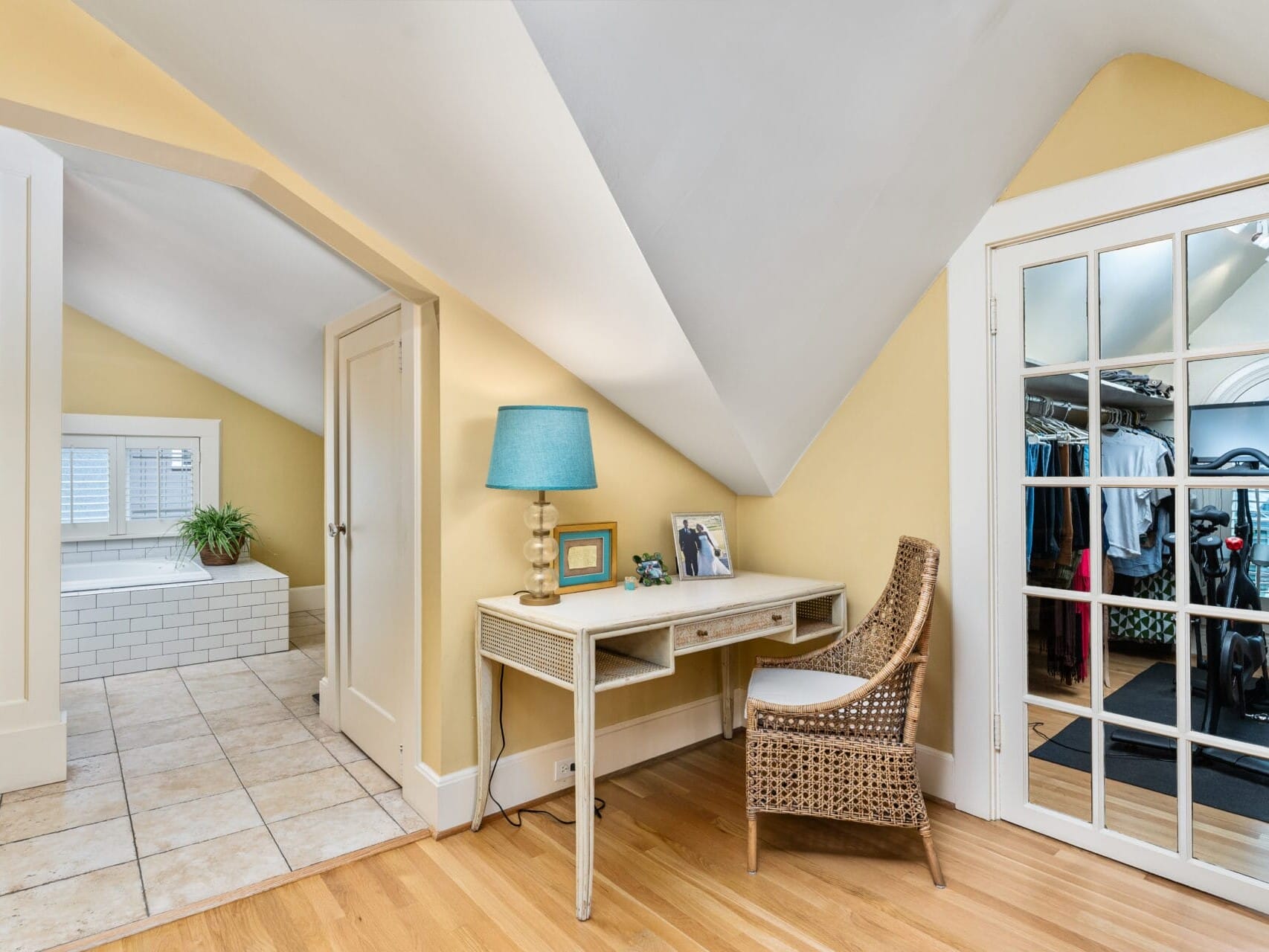 Bright attic room featuring a small desk with a wicker chair and a turquoise lamp. A door with glass panes opens to a walk-in closet. An open door reveals a bathroom with a white tiled bathtub. Walls are painted yellow, with light wood flooring.