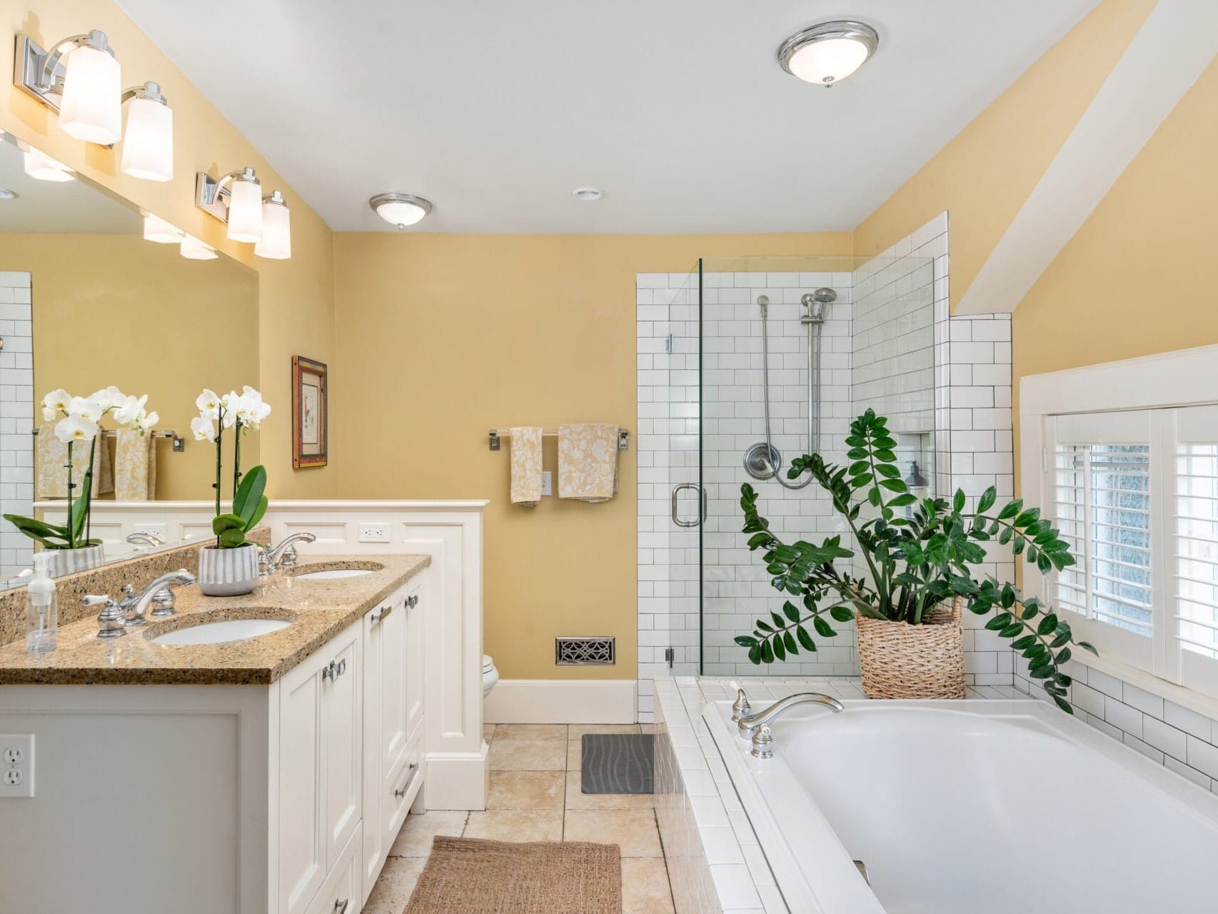 A bright bathroom with yellow walls featuring a double sink vanity with a granite countertop and mirror, a potted plant by a soaking tub, and a glass-enclosed shower. The floor is tiled, and natural light streams through the shuttered window.