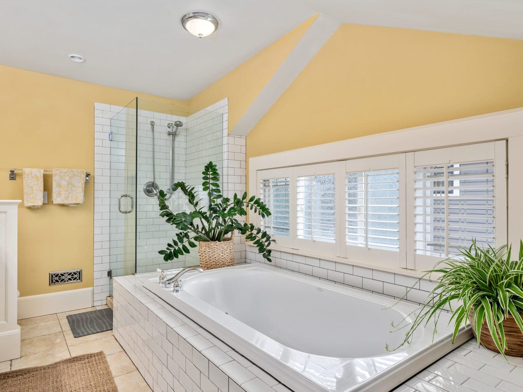 A bathroom with a white bathtub surrounded by indoor plants, a glass-enclosed shower, and off-white tiled walls. Yellow walls and light wooden blinds add warmth to the space. Two patterned towels hang on the left wall.