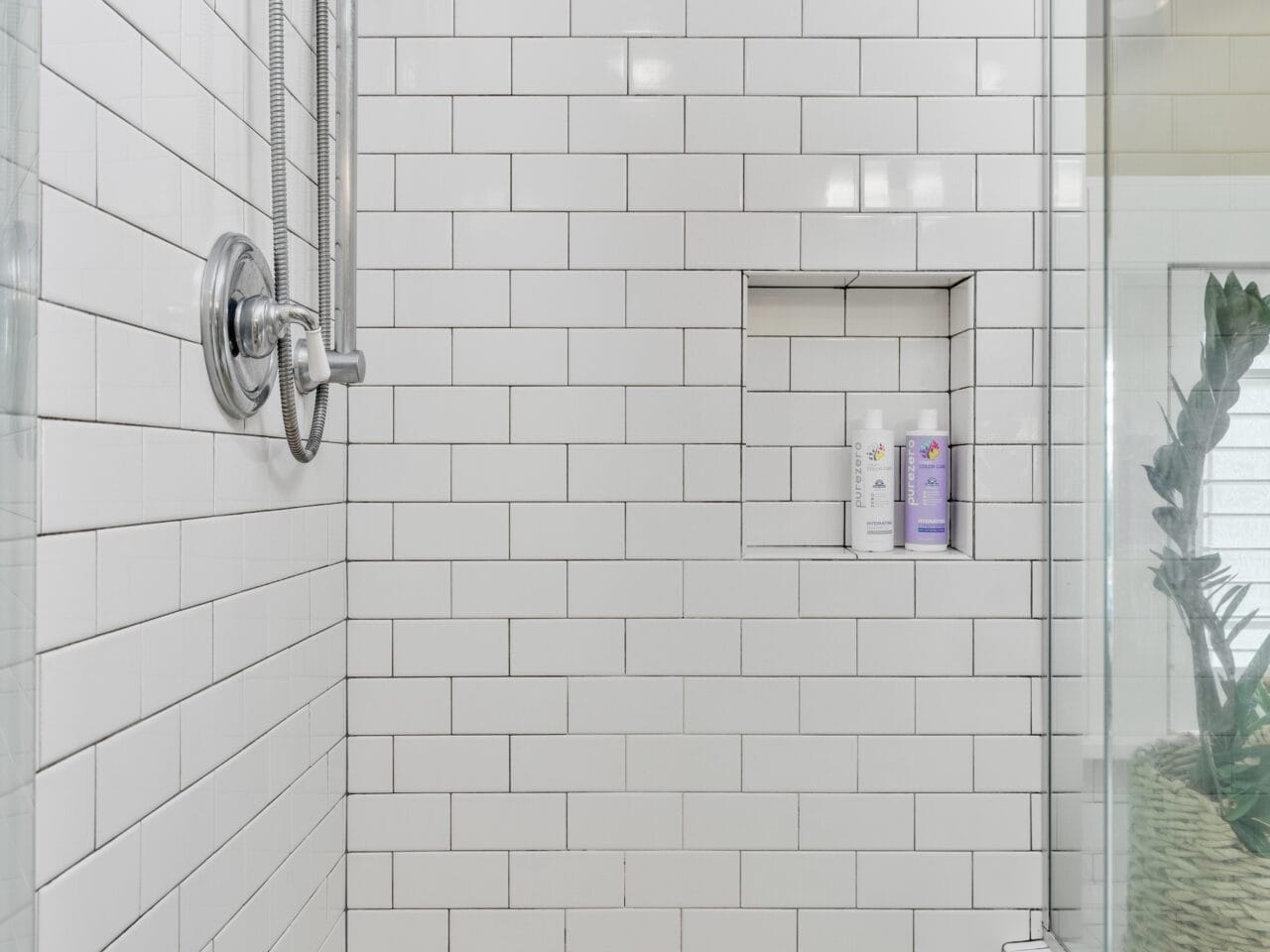 A shower with white subway tiles features a stainless steel showerhead and handle. There is a built-in niche holding toiletries. The floor is a light brown speckled surface, and a glass panel on the right with a plant visible behind it.