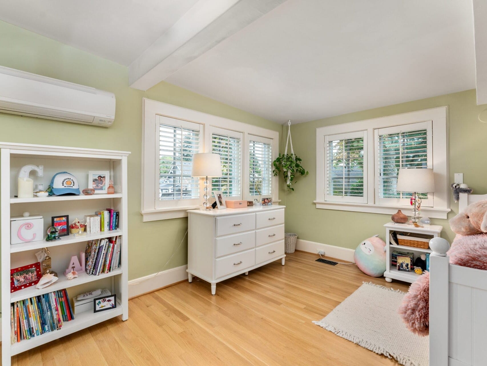 A cozy childrens bedroom with light green walls, featuring a white bookshelf filled with toys and books, a white dresser with a lamp and decorations, a hanging plant near the window, and a bed with fluffy pillows on the right.