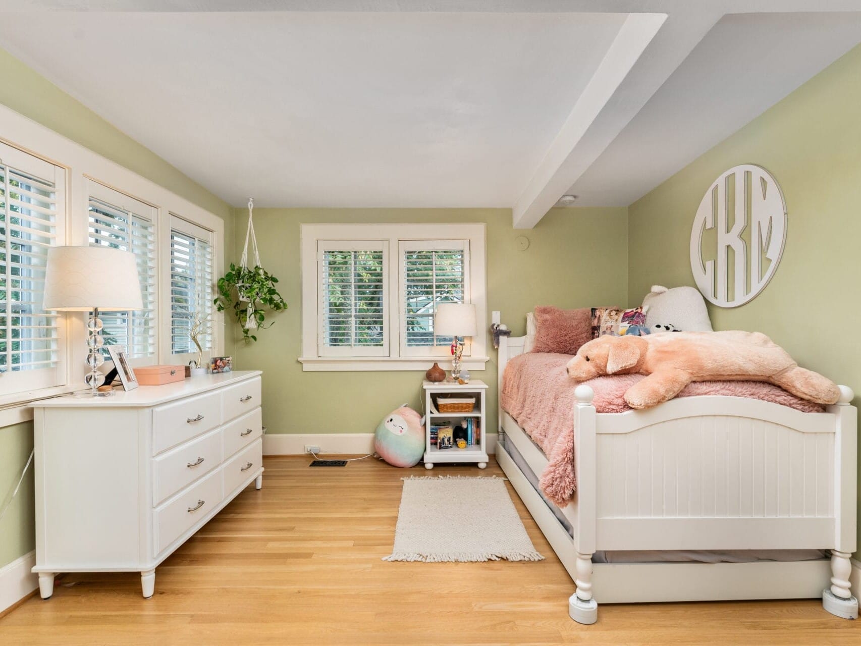 Bright, cozy bedroom with green walls, a bed adorned with pink pillows and a large teddy bear. A white dresser with a lamp and plants sits by the window. Hardwood floors and natural light from the shuttered windows create a warm atmosphere.