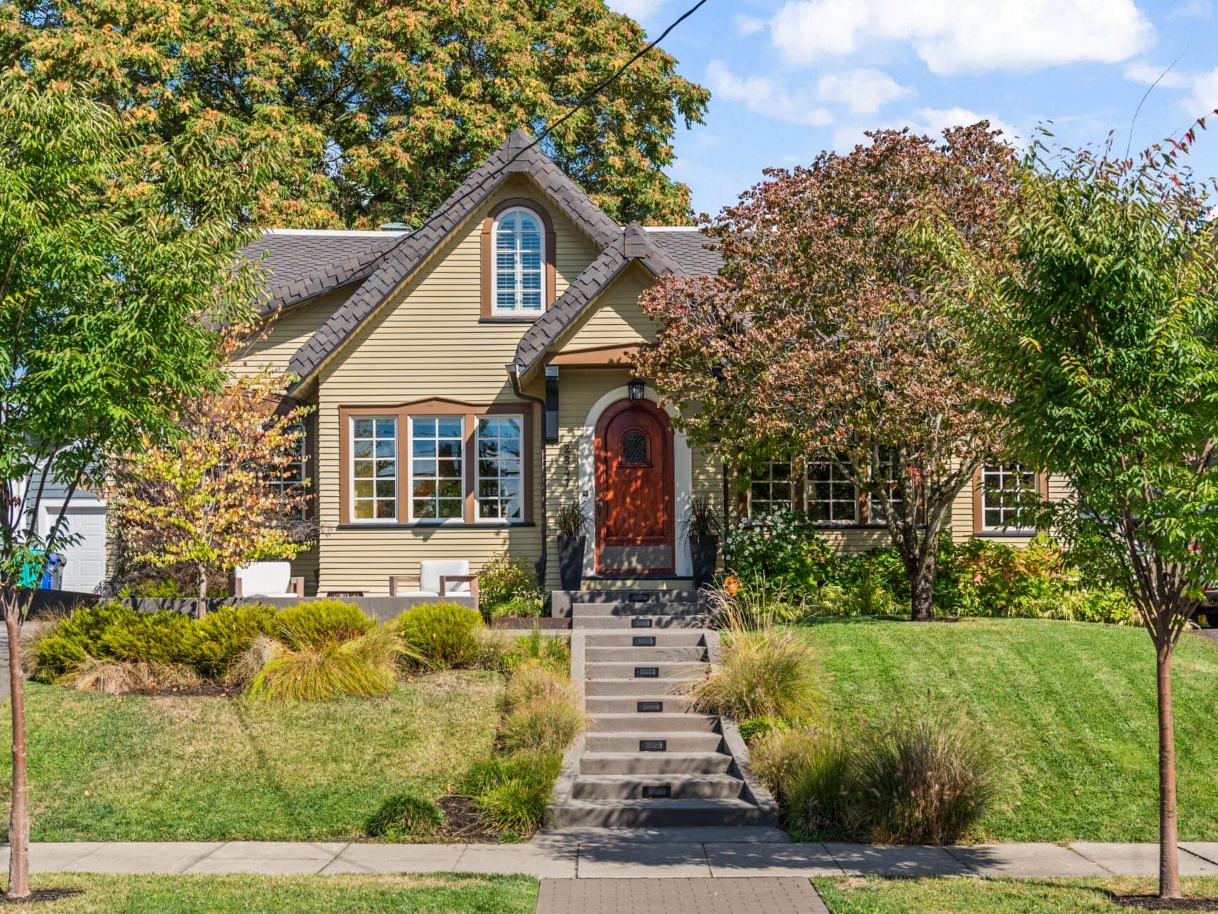 A charming house with a beige exterior and red front door, surrounded by lush green trees and well-maintained landscaping. Steps lead up from the sidewalk, and a large tree shades the left side of the lawn.