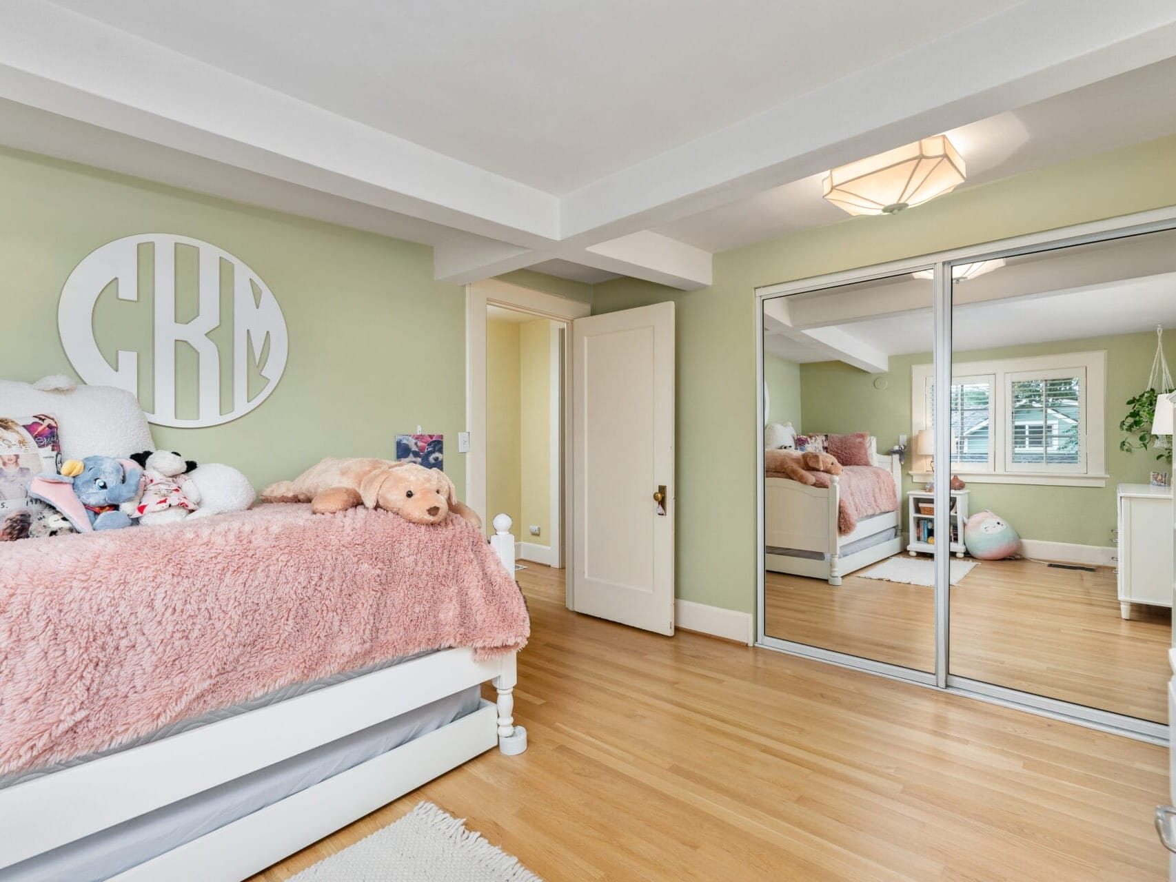 A cozy bedroom with a twin bed featuring pink bedding and plush toys. The walls are light green with a circular decor piece. A mirrored wardrobe reflects the space, and a window allows natural light. A soft rug lies on the hardwood floor.