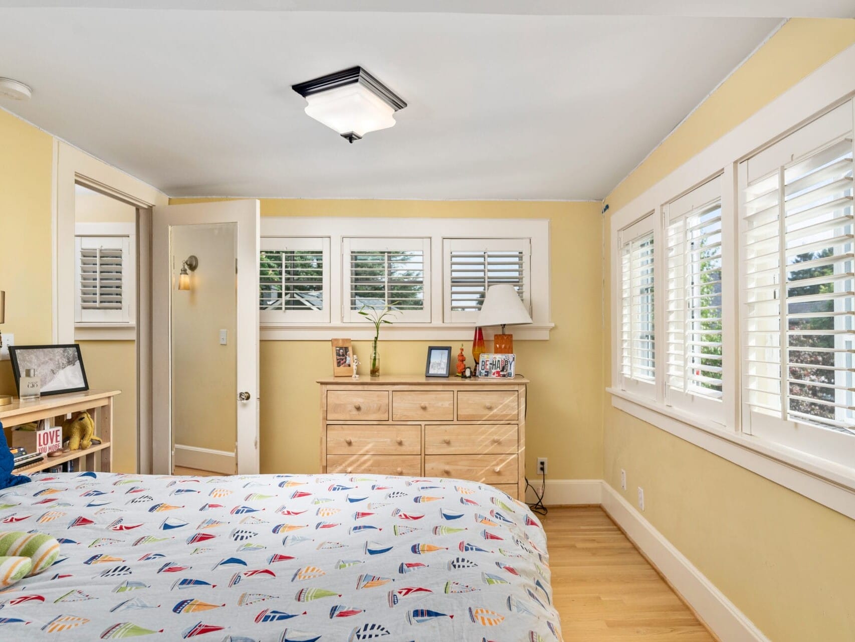 A cozy bedroom with yellow walls and wooden floor features a bed with a colorful sailboat-patterned duvet. A wooden dresser displays framed photos and decor. White shutters cover large windows, allowing natural light to fill the room.