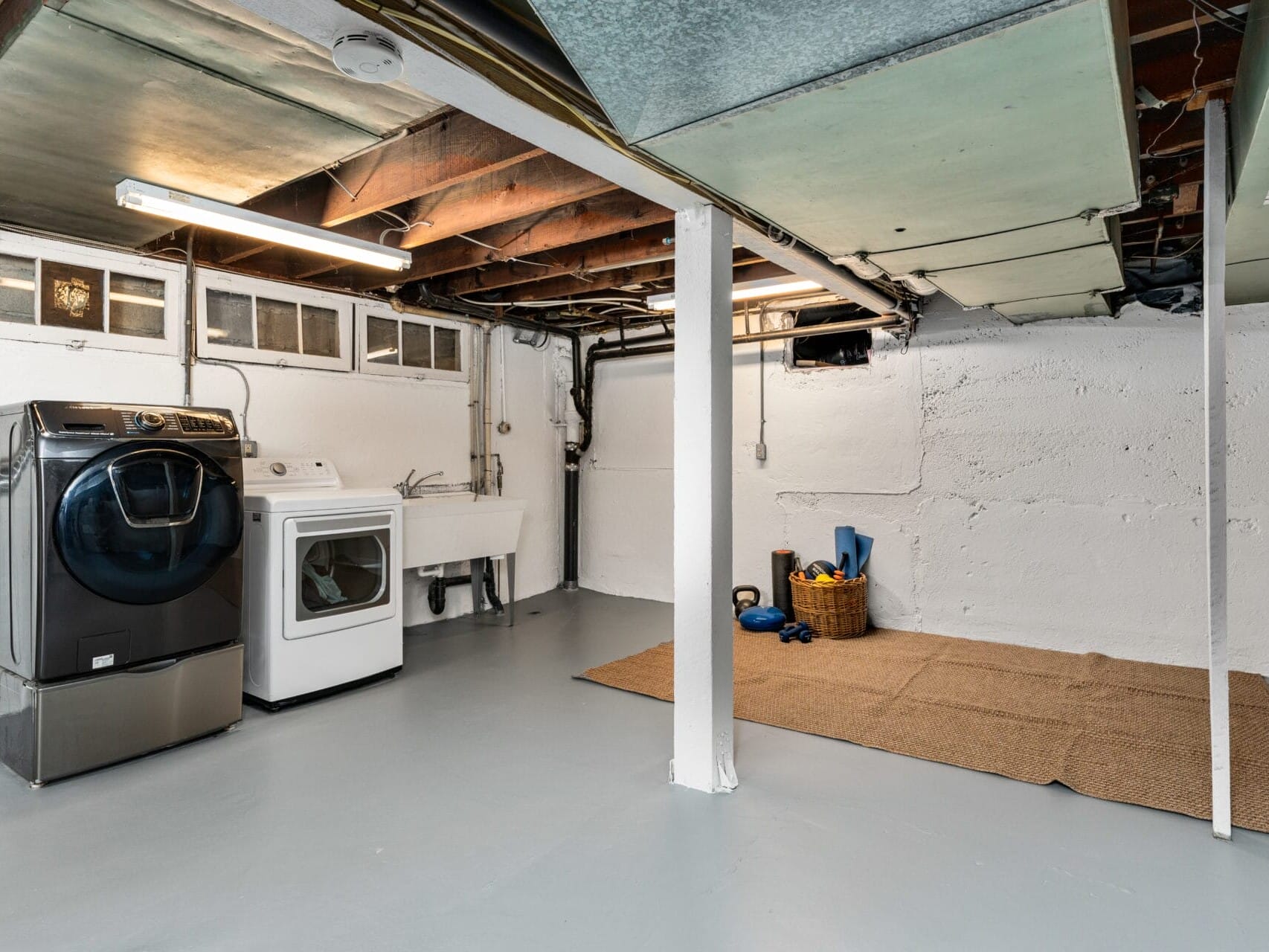 Basement room with a washing machine, dryer, and sink on one side. On the other side, a yoga mat and a basket with exercise equipment are placed on a mat. The ceiling has exposed beams and ducts. Walls are painted white and floor is gray.