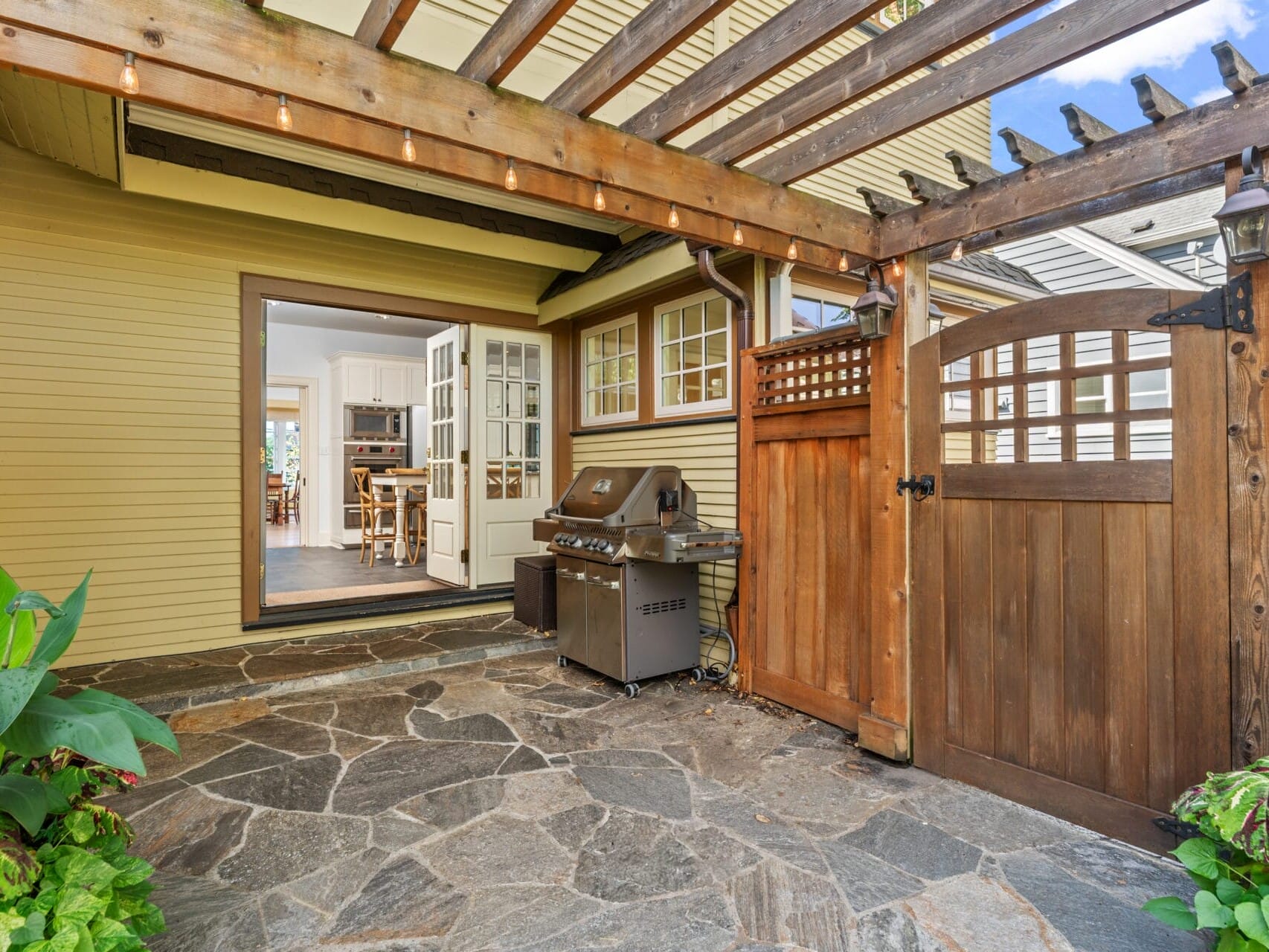 A cozy patio with stone flooring, a wooden pergola, and a barbecue grill. The area leads to a kitchen through open French doors. The patio is adorned with potted plants and a wooden gate, creating a warm, inviting atmosphere.