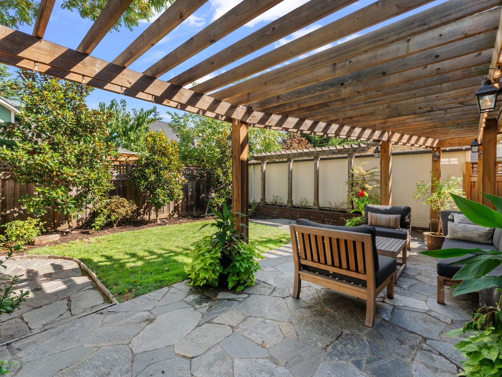 A cozy backyard patio with a wooden pergola and stone flooring. Wooden furniture with cushions sits under the pergola, surrounded by lush plants and trees. Sunlight filters through the slats, creating a warm, inviting atmosphere.