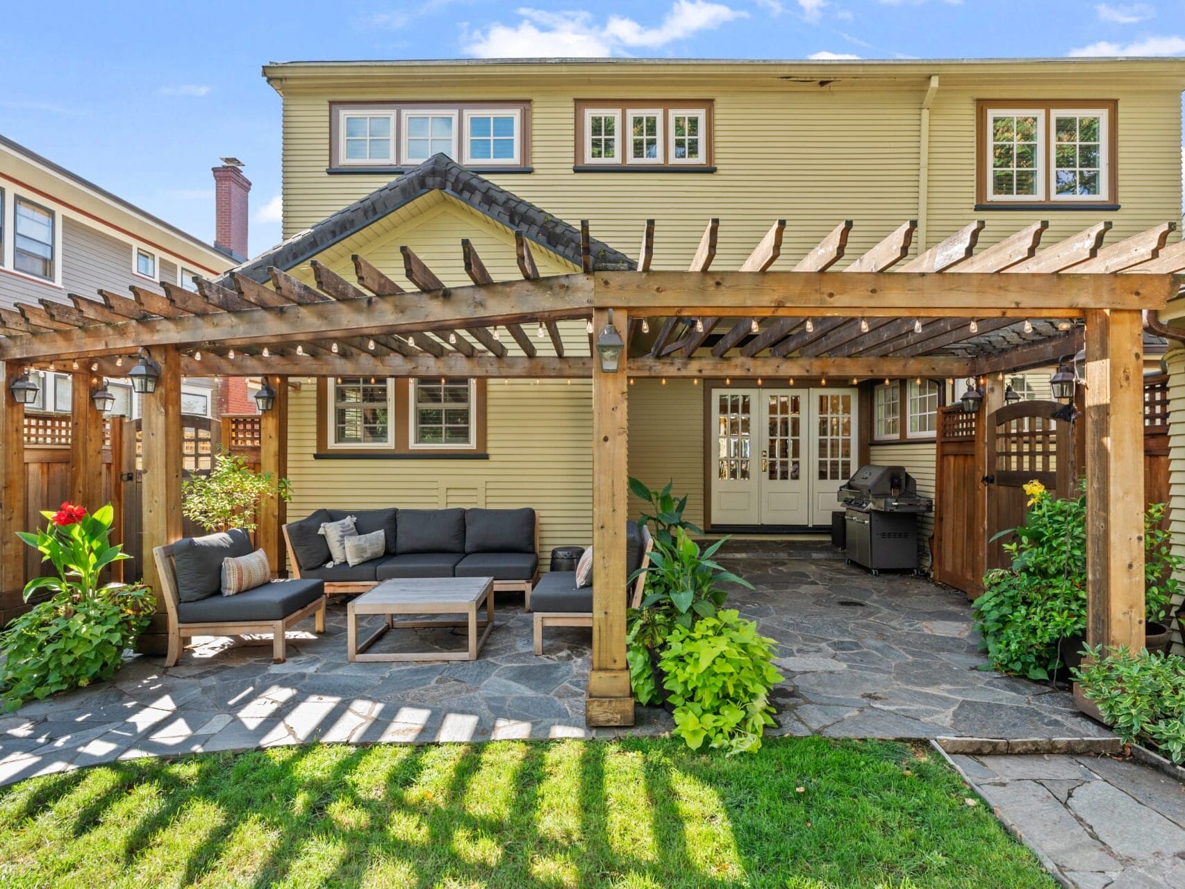 A backyard features a wooden pergola covering a patio with a sofa, chairs, and a coffee table. A grill stands in the corner. Lush plants surround the area, and a two-story house is visible in the background.