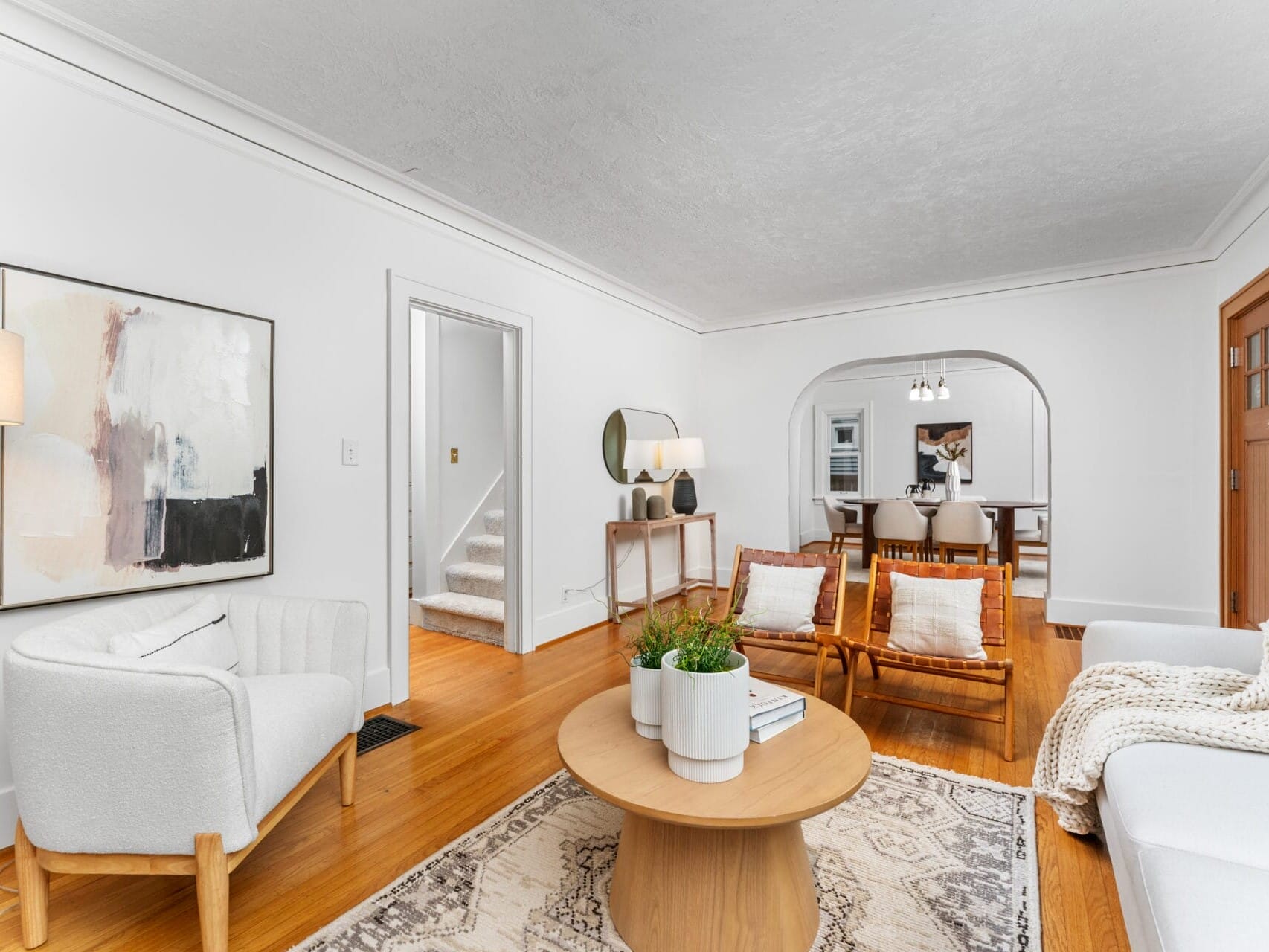 A cozy living room with white walls, featuring a large abstract painting, a soft armchair, and a wooden coffee table with plants. There are woven chairs, a patterned rug, and an arched opening leading to a dining area in the background.