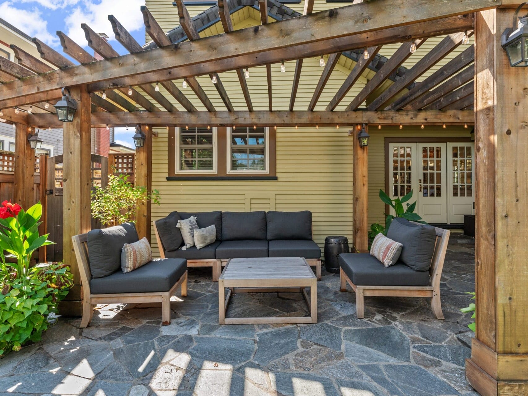 A wooden pergola shelters a cozy outdoor seating area with dark gray cushions and a rustic wooden coffee table. Potted plants and red flowers add a touch of color to the stone patio, while a yellow house with double French doors is in the background.