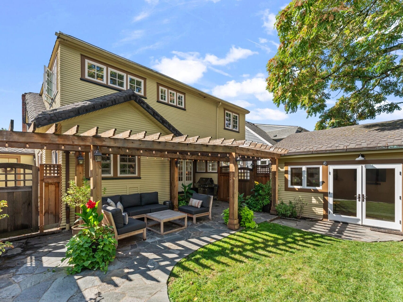 A backyard patio featuring a wooden pergola with seating area, surrounded by lush greenery. Adjacent is a tan house with multiple windows and a smaller structure with white double doors. The sky above is clear and blue.