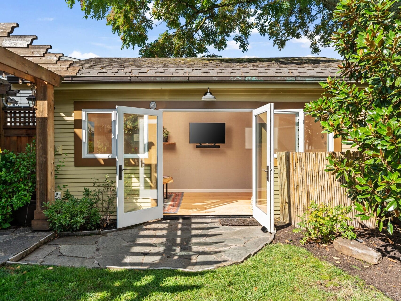 Open double doors lead to a cozy room with a wall-mounted TV and wooden flooring. The exterior features a pergola, stone path, lush greenery, and a bamboo privacy screen. A tree provides shade, creating a serene backyard atmosphere.