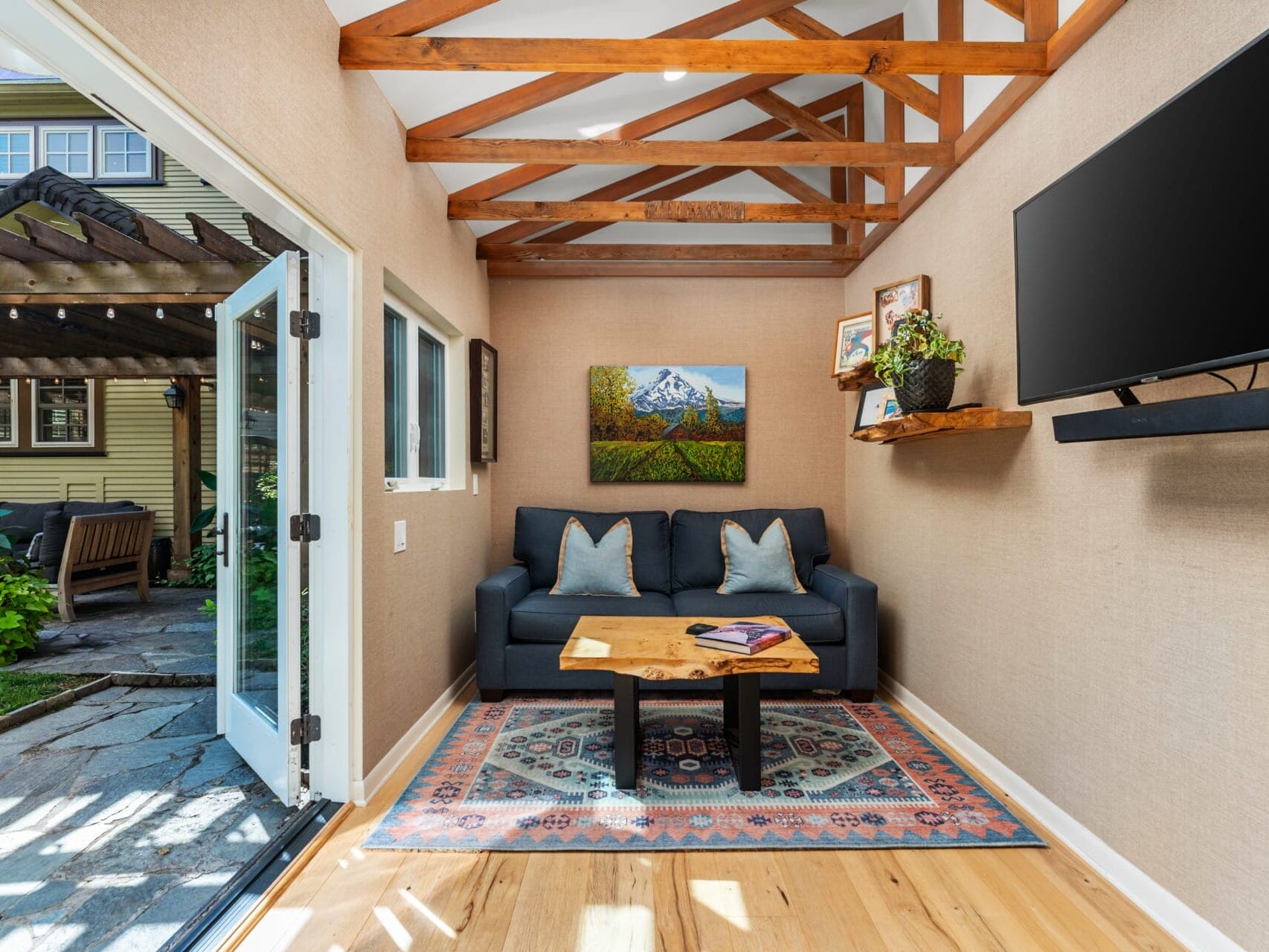 A cozy room with a blue sofa, patterned rug, and a coffee table. A TV is mounted on the wall with a shelf beneath, displaying plants and framed photos. Large open doors lead to a patio with wooden pergola and greenery. Ceiling has exposed wooden beams.