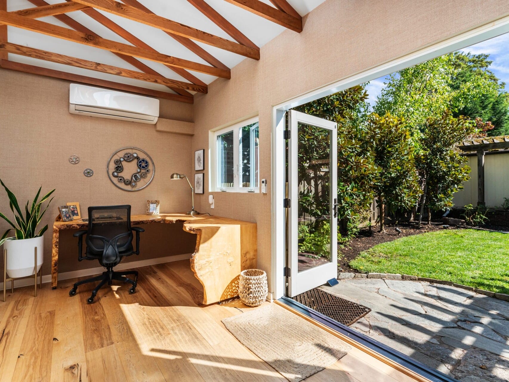 A bright home office with wooden floors and exposed beams features an airy, open door to a lush garden. A rustic wooden desk holds a black chair, small decorations, and a potted plant. The space is well-lit and inviting.