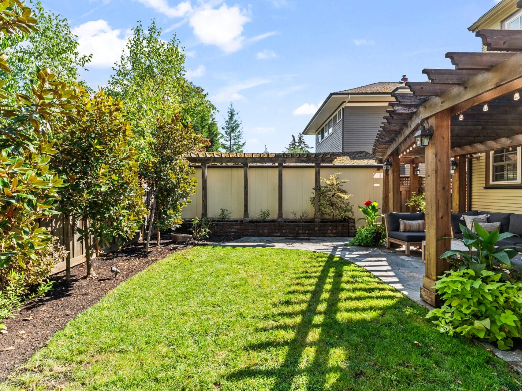 A backyard garden with a grass lawn, surrounded by a wooden fence and trees. A patio with cushioned seating is shaded by a wooden pergola. String lights hang from the pergola, creating a cozy atmosphere. Sky is clear and sunny.