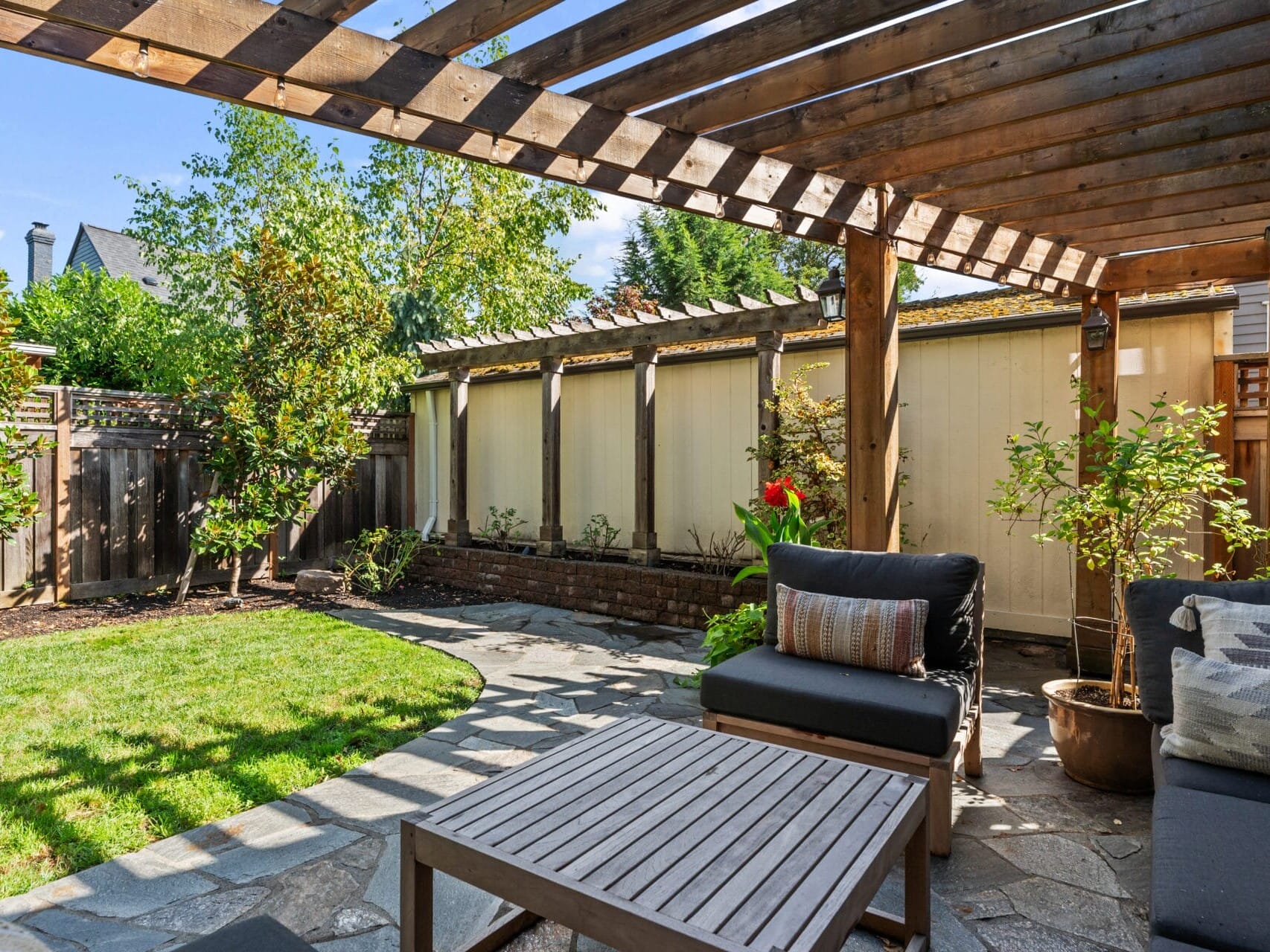 A backyard patio with a wooden pergola, outdoor seating, and a small table. The area features potted plants and overlooks a green lawn, enclosed by a wooden fence. Sunlight illuminates the scene, creating a cozy and inviting atmosphere.