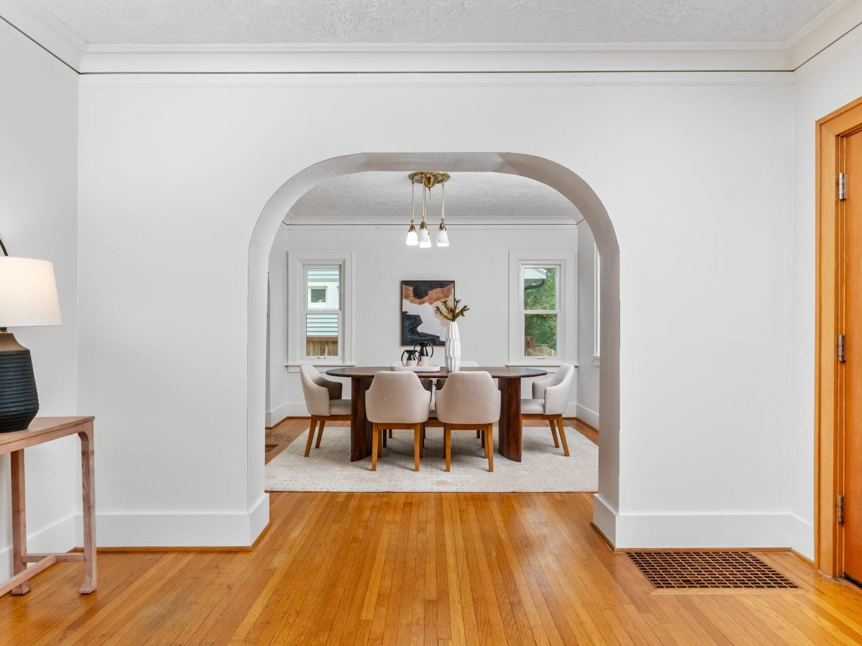 A bright dining room with a wooden floor, white walls, and a wooden table surrounded by light-colored chairs. Theres an archway leading to the dining area and a side table with a lamp on the left. A wooden front door is visible on the right.