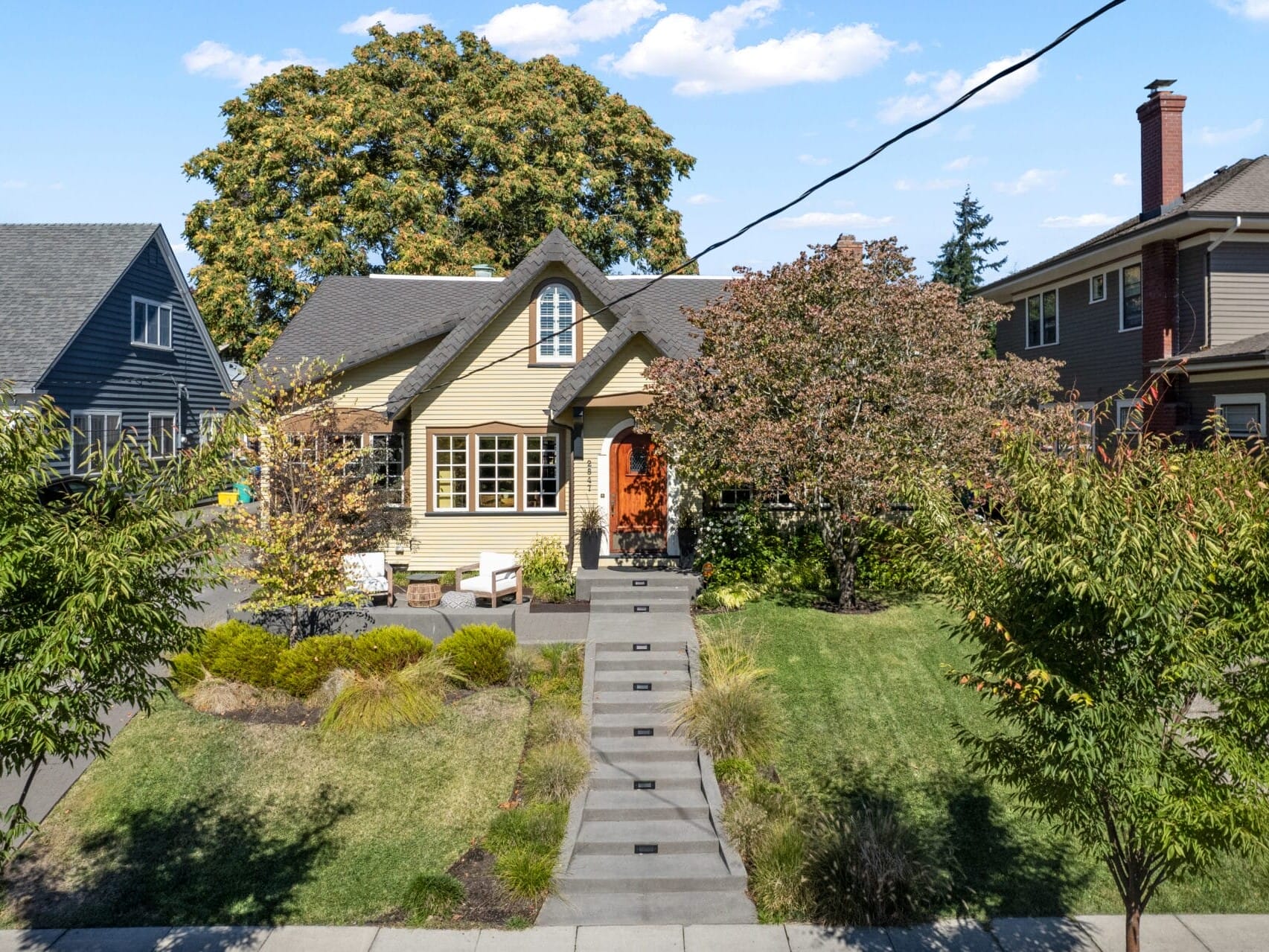 A charming suburban house with a steep, landscaped front yard and a central staircase leading to the entrance. The house has a peaked roof, trees, and shrubs in the garden. Neighboring houses frame the scene under a blue sky.