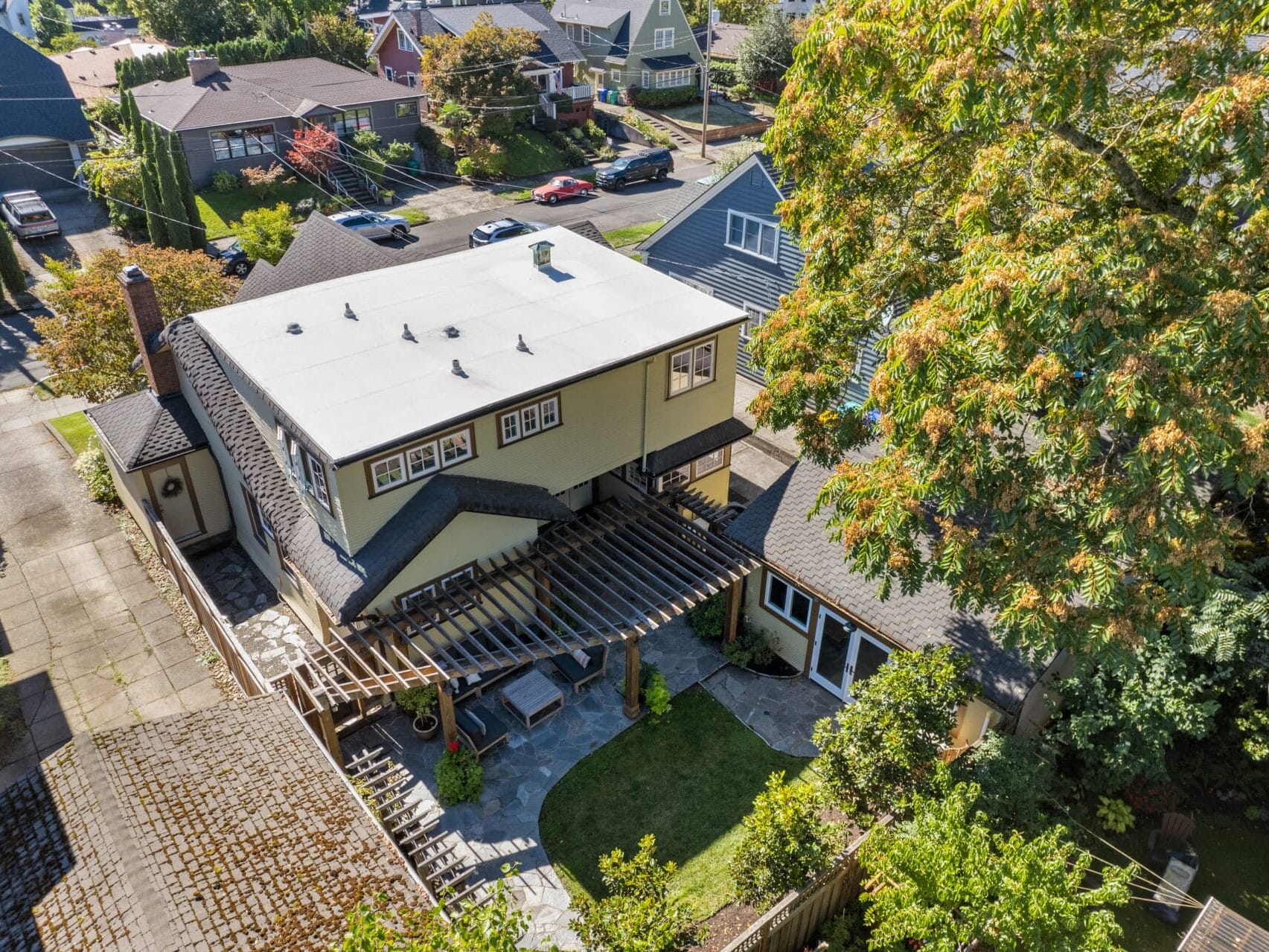 Aerial view of a suburban neighborhood featuring a beige two-story house with a flat roof and pergola in the backyard. The yard includes a small lawn, trees, and a paved patio. Nearby houses line a street in the background.