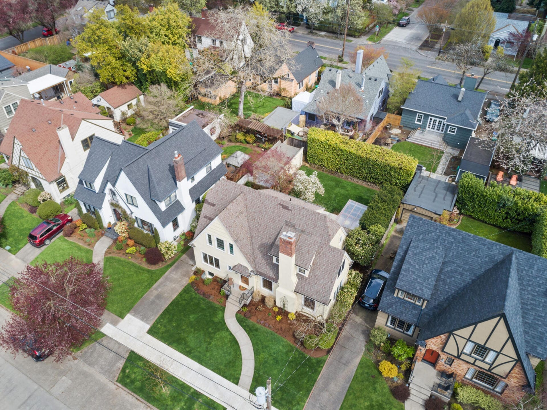 Aerial view of a suburban neighborhood featuring several houses. Each house has a neatly landscaped garden with green lawns and trees. The street is lined with parked cars, and the scene is surrounded by roads and more greenery.