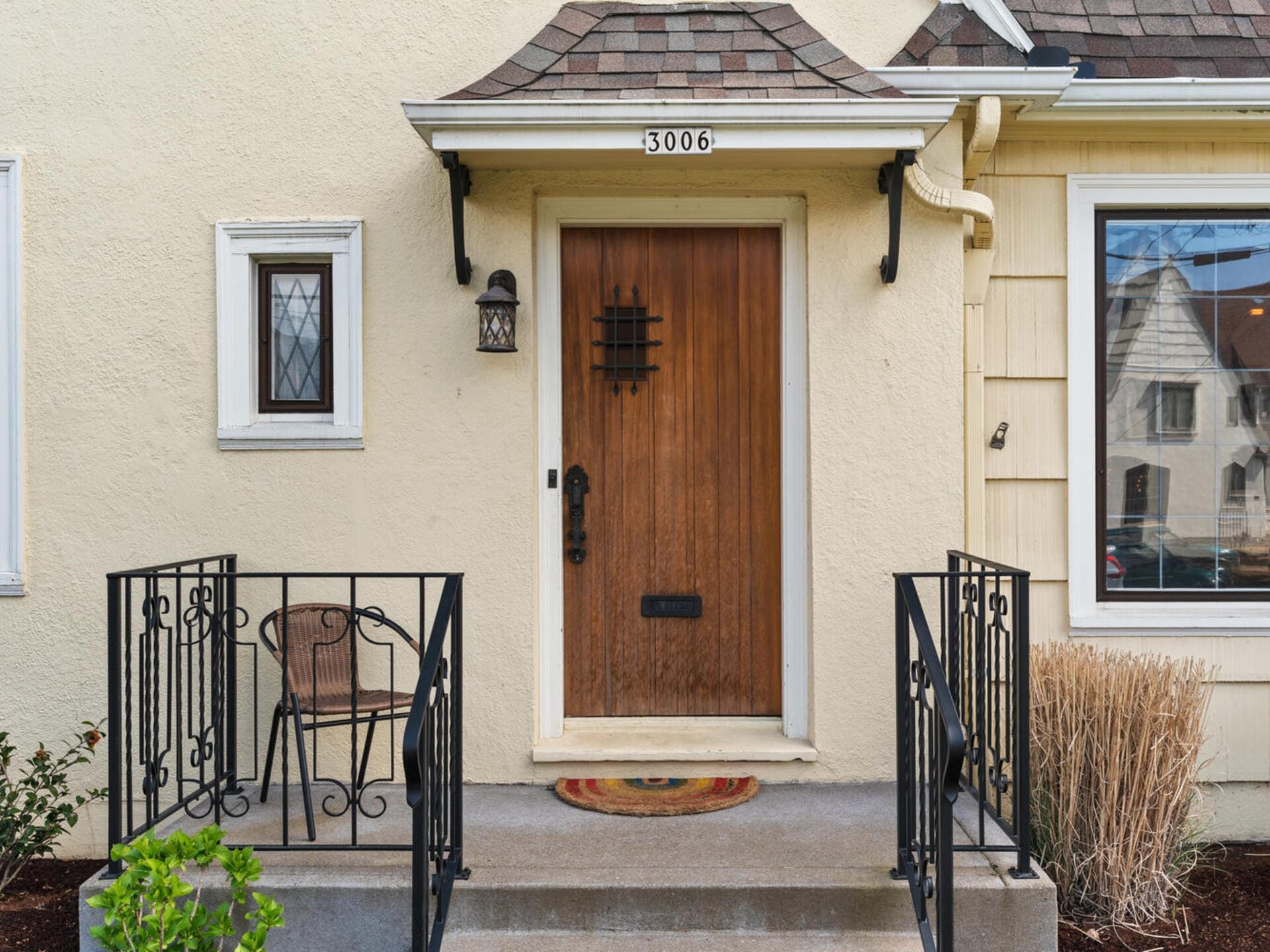 A house entrance featuring a wooden door with the number 3006 above. The door is flanked by a small window and a larger window. Black wrought iron railings border the concrete steps leading to the door. A wicker chair sits to the left.