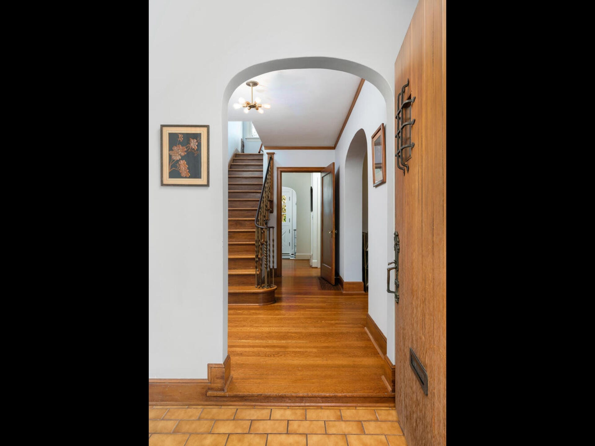 Interior of a home featuring a wooden staircase with ornate railing, leading to an upper floor. The entryway has wood flooring and white walls, adorned with framed art. Wooden door is partially open, revealing a warmly lit hallway.