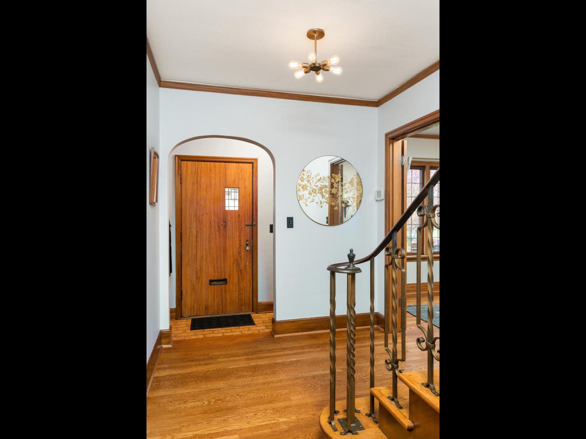 A hallway with a wooden front door featuring a mail slot and window, a circular mirror on the wall, and a staircase with ornate metal railings. A small chandelier hangs from the ceiling, and sunlight filters in from a window on the right.
