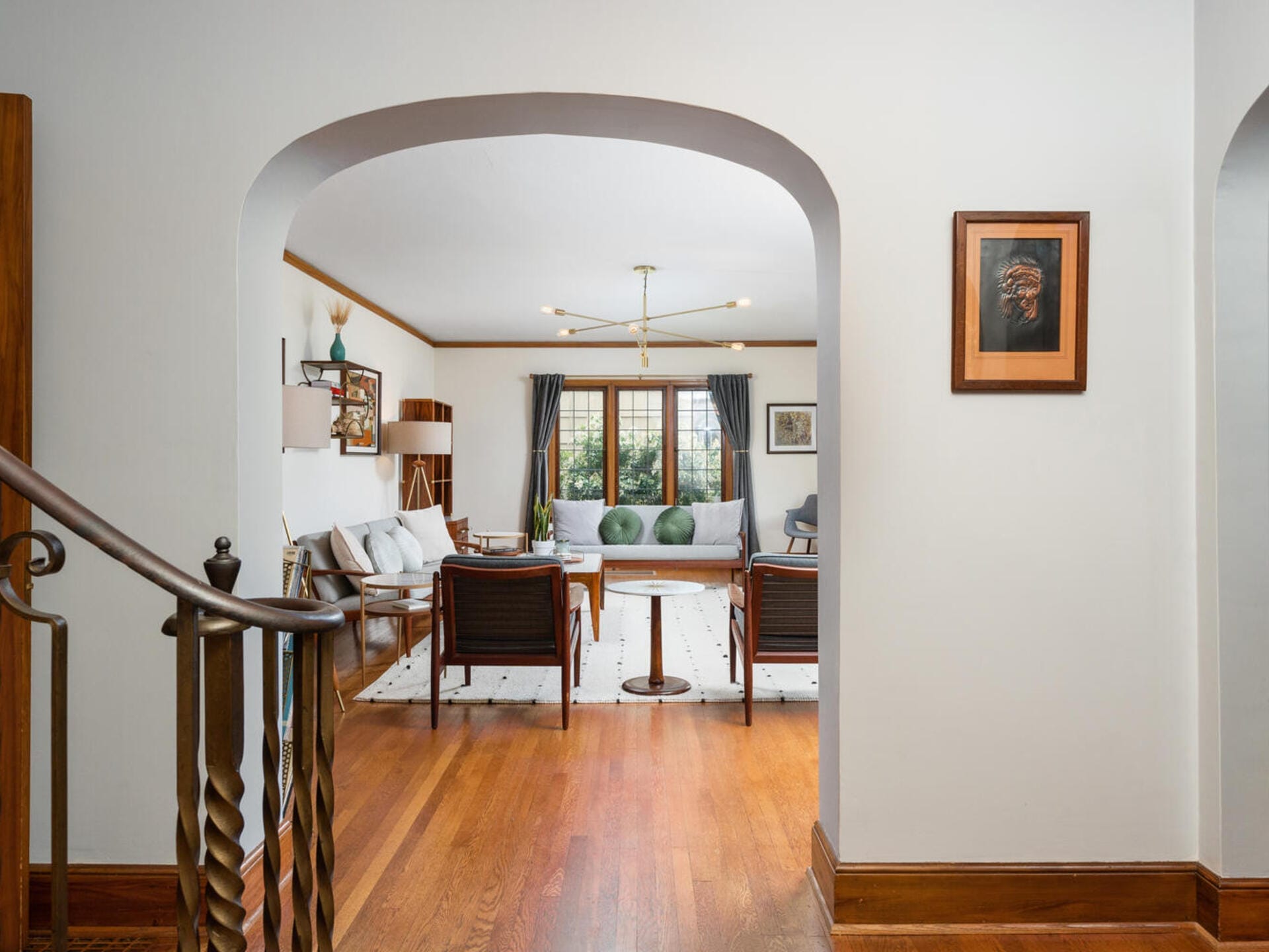 A spacious living room with wooden floors, white walls, and a large archway entrance. It features a modern chandelier, mid-century furniture, and large windows revealing an outdoor view. A picture hangs on the wall by the staircase.