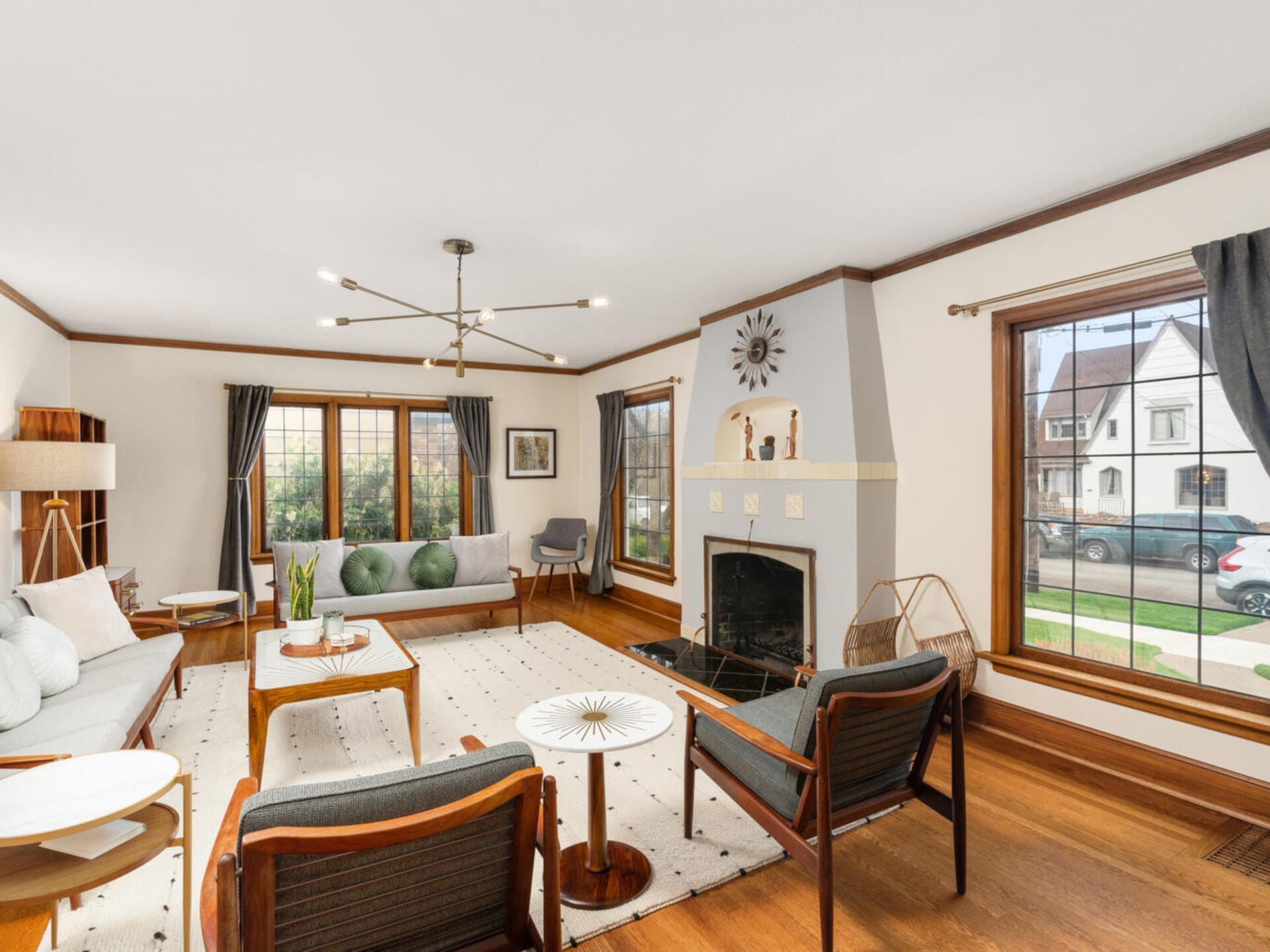 A spacious living room with wooden floors, featuring a modern chandelier, a large rug, and a gray fireplace. It has a sofa, armchairs, and a round coffee table. Large windows with dark curtains let in natural light, offering a view of houses outside.