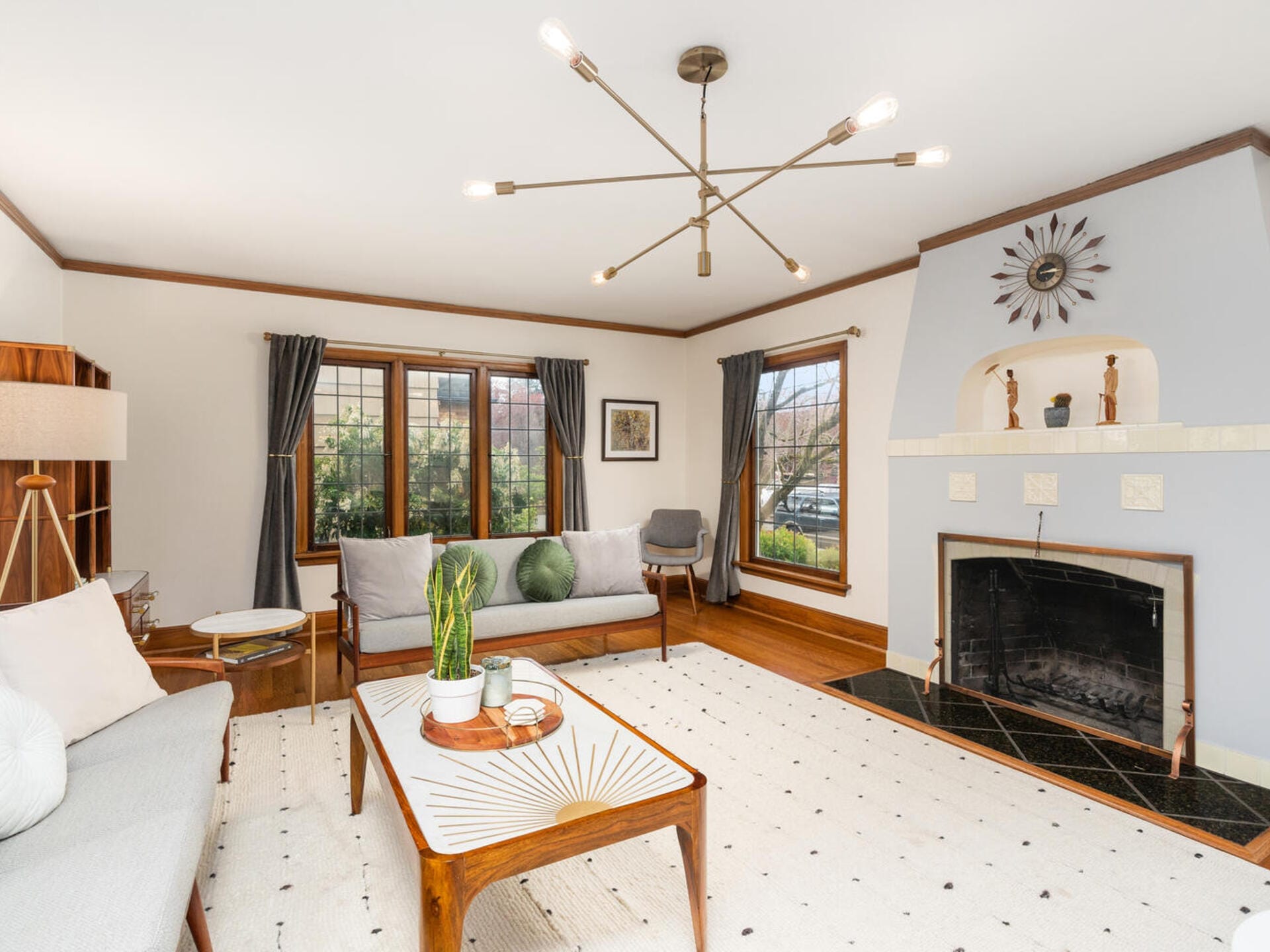 A stylish living room with mid-century modern decor, featuring a gray sofa, wooden coffee table, and a black fireplace. The room has large windows with dark curtains, a geometric light fixture, and light-colored walls and rug.