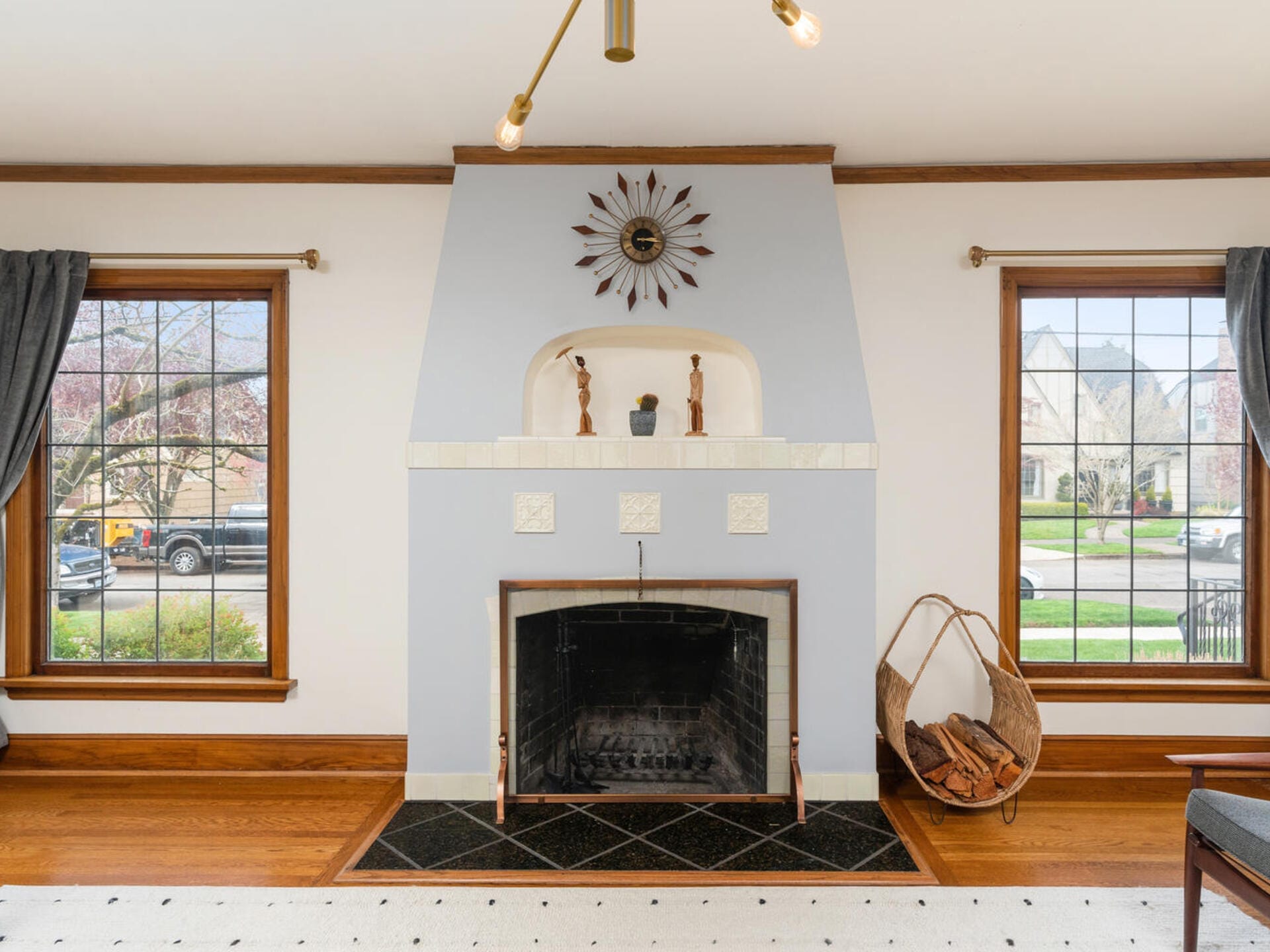 A cozy living room features a symmetrical fireplace with a decorative clock above it. Two windows with dark curtains flank the fireplace, and a wicker basket filled with firewood sits to the right. The room has hardwood floors and light gray walls.