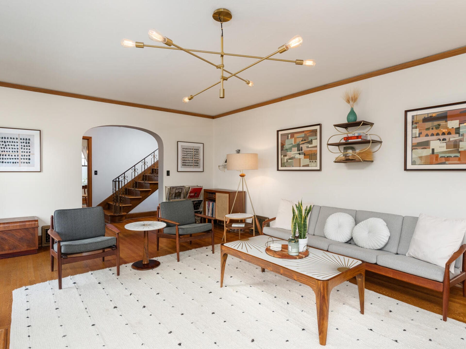 A modern living room with a minimalist design, featuring a gray sofa, two matching chairs, a coffee table, and a geometric light fixture. The room has wooden floors, framed art on the walls, and a white textured rug. A staircase is visible in the background.