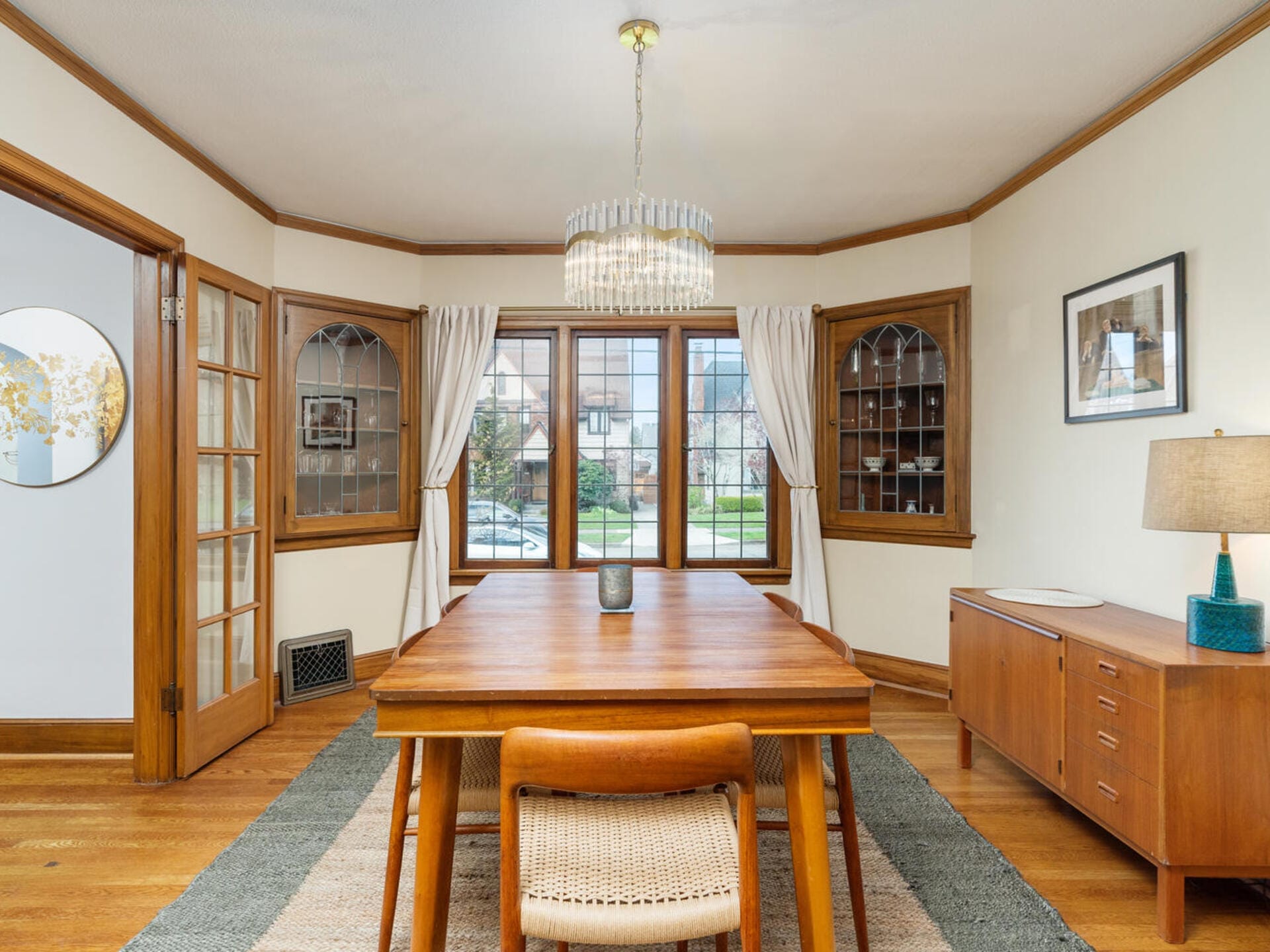 A dining room featuring a wooden table and chairs, a sideboard with a lamp, and large windows with white curtains. The chandelier overhead adds elegance. The walls are adorned with artwork, and built-in cabinets are visible in the corners.