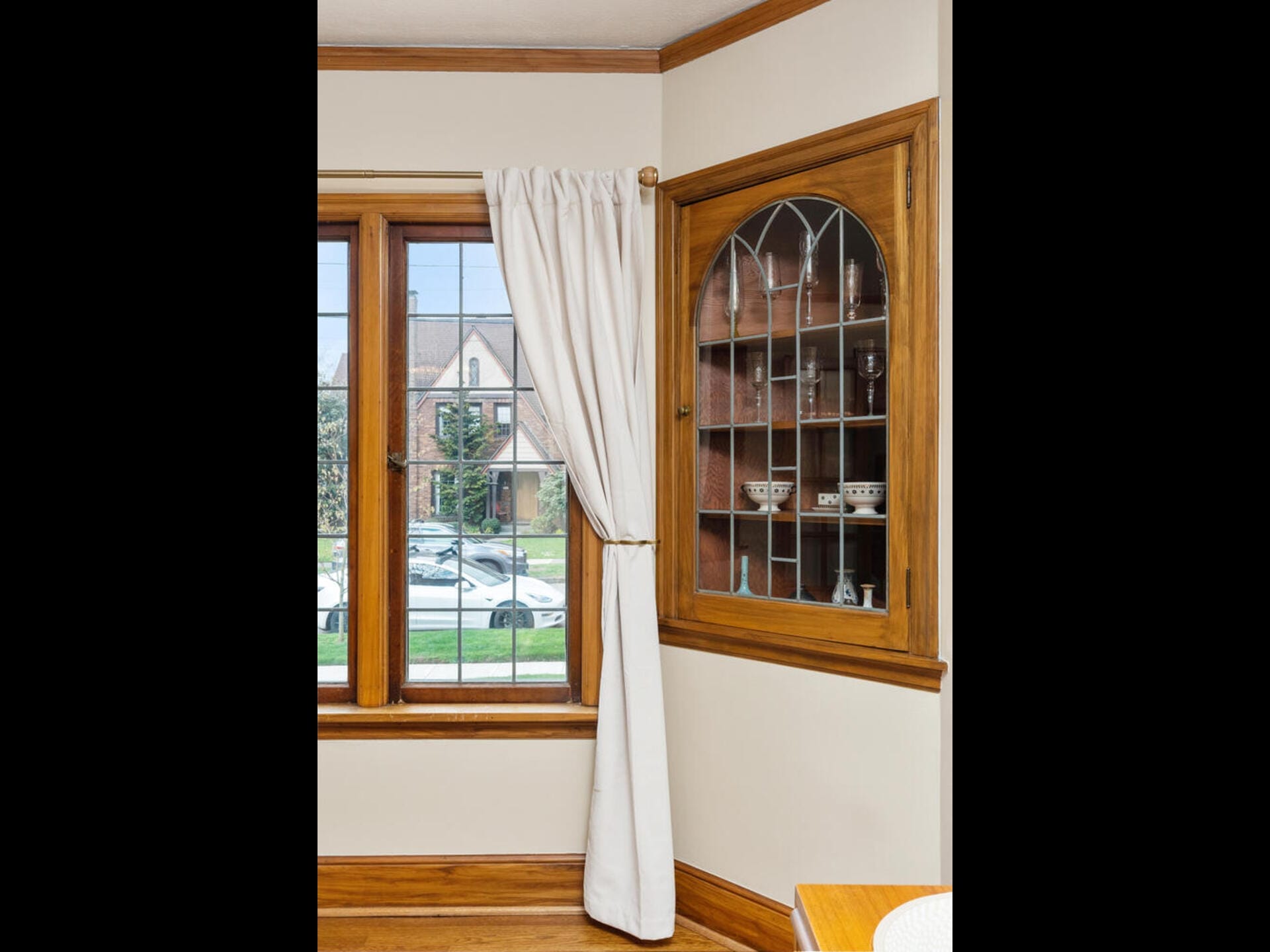 Interior of a room with a large wooden-framed window partially covered by white curtains. Next to it is a built-in cabinet with glass doors displaying small decorative items. Outside, a house and greenery are visible.