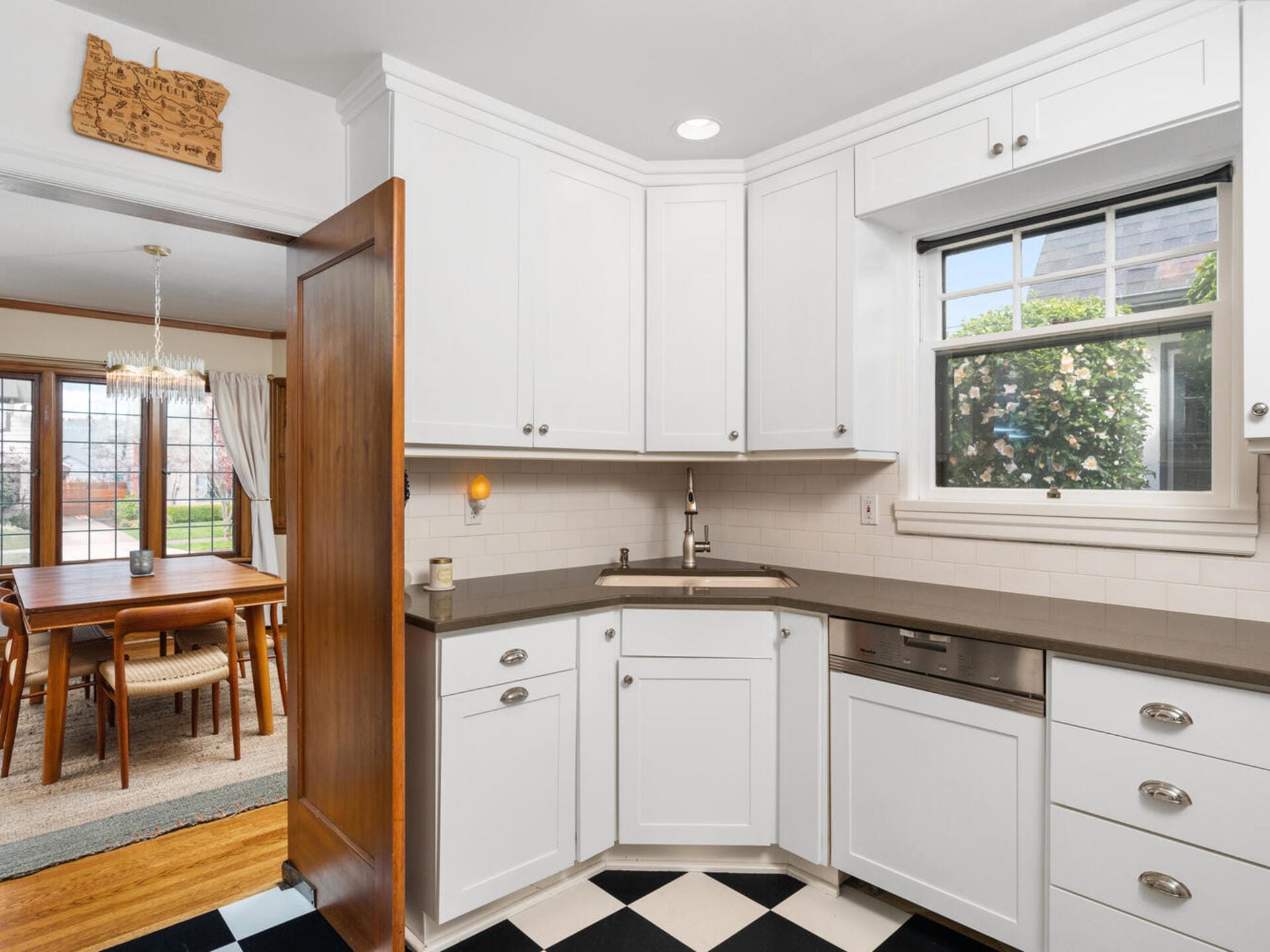 Bright kitchen with white cabinetry, gray countertops, and a stainless steel sink below a window. The floor features a black and white checkered pattern. An open door leads to a dining area with a wooden table and chairs, visible through the doorway.