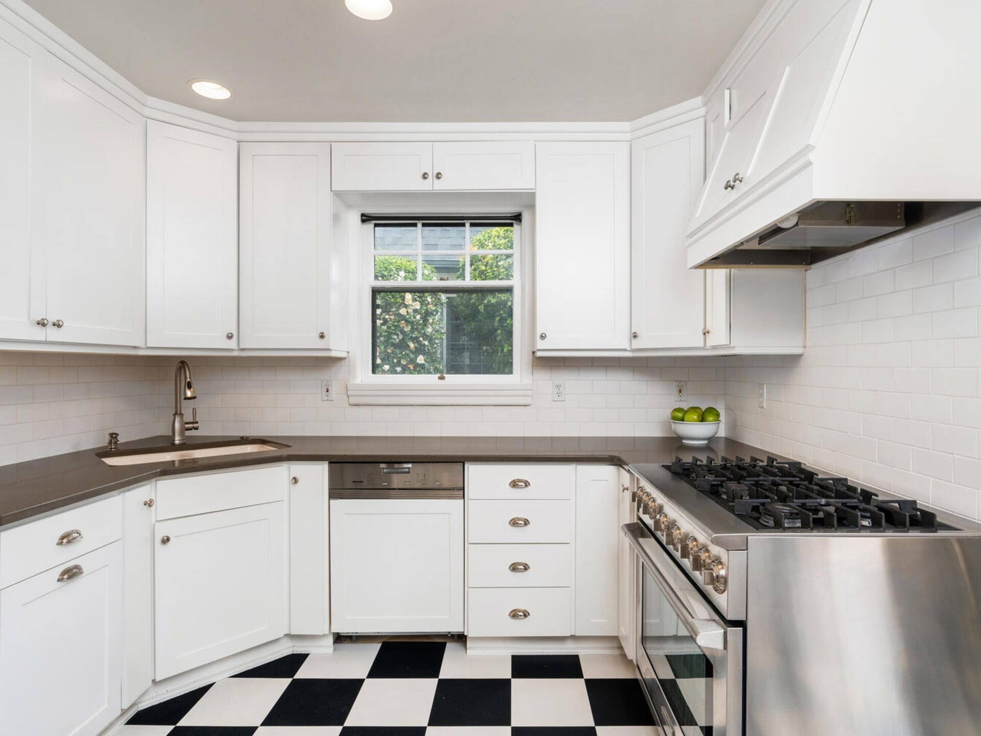 A modern kitchen with white cabinetry, a stainless steel stove, and a dishwasher. The countertops are dark, and there is a window above the sink. The floor features a black and white checkerboard pattern.