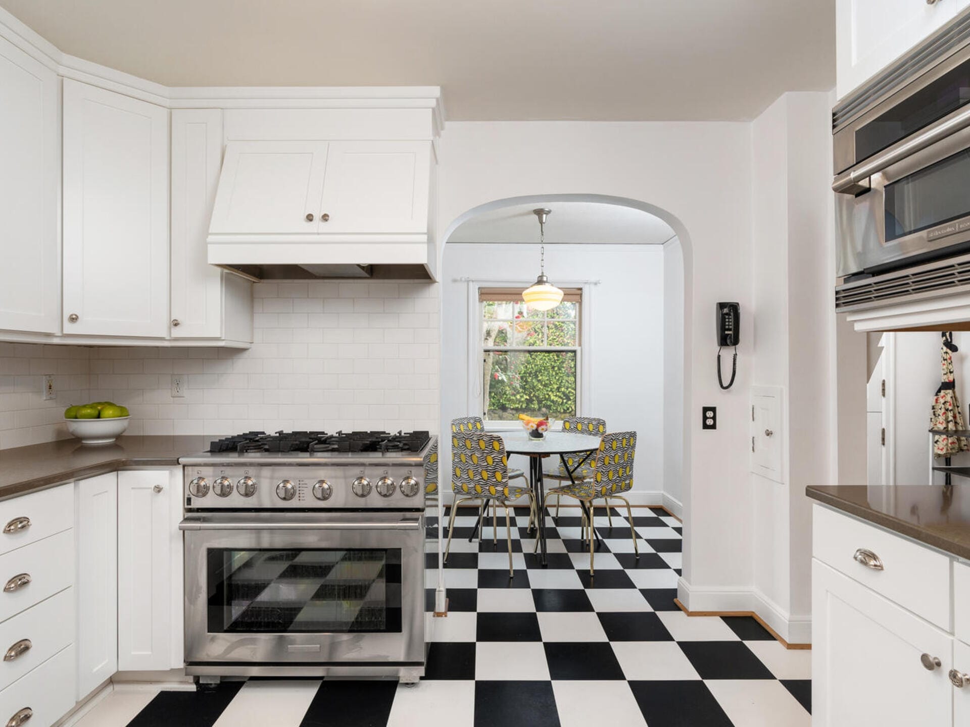 A modern kitchen with white cabinets and a black-and-white checkered floor. A stainless steel stove and oven are on the left. Through an archway, a dining area with a round table and patterned chairs is visible. A window shows greenery outside.