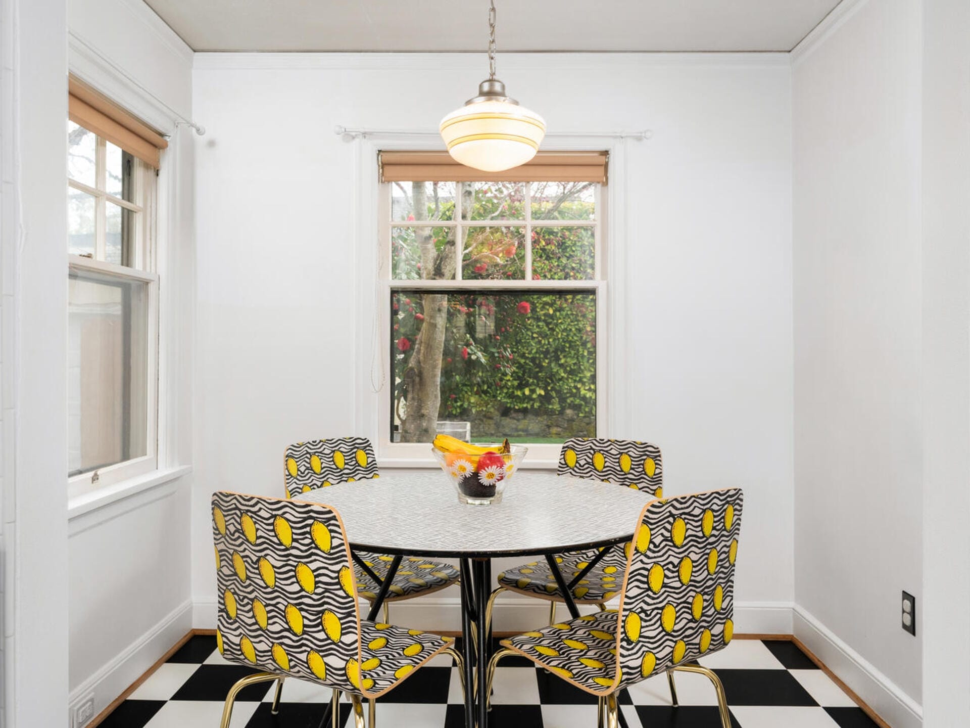 A dining area with a round white table and four patterned chairs. A bowl of fruit and flowers sits on the table. The space is surrounded by white walls, with two windows offering a view of greenery outside. Black and white checkered flooring adds contrast.