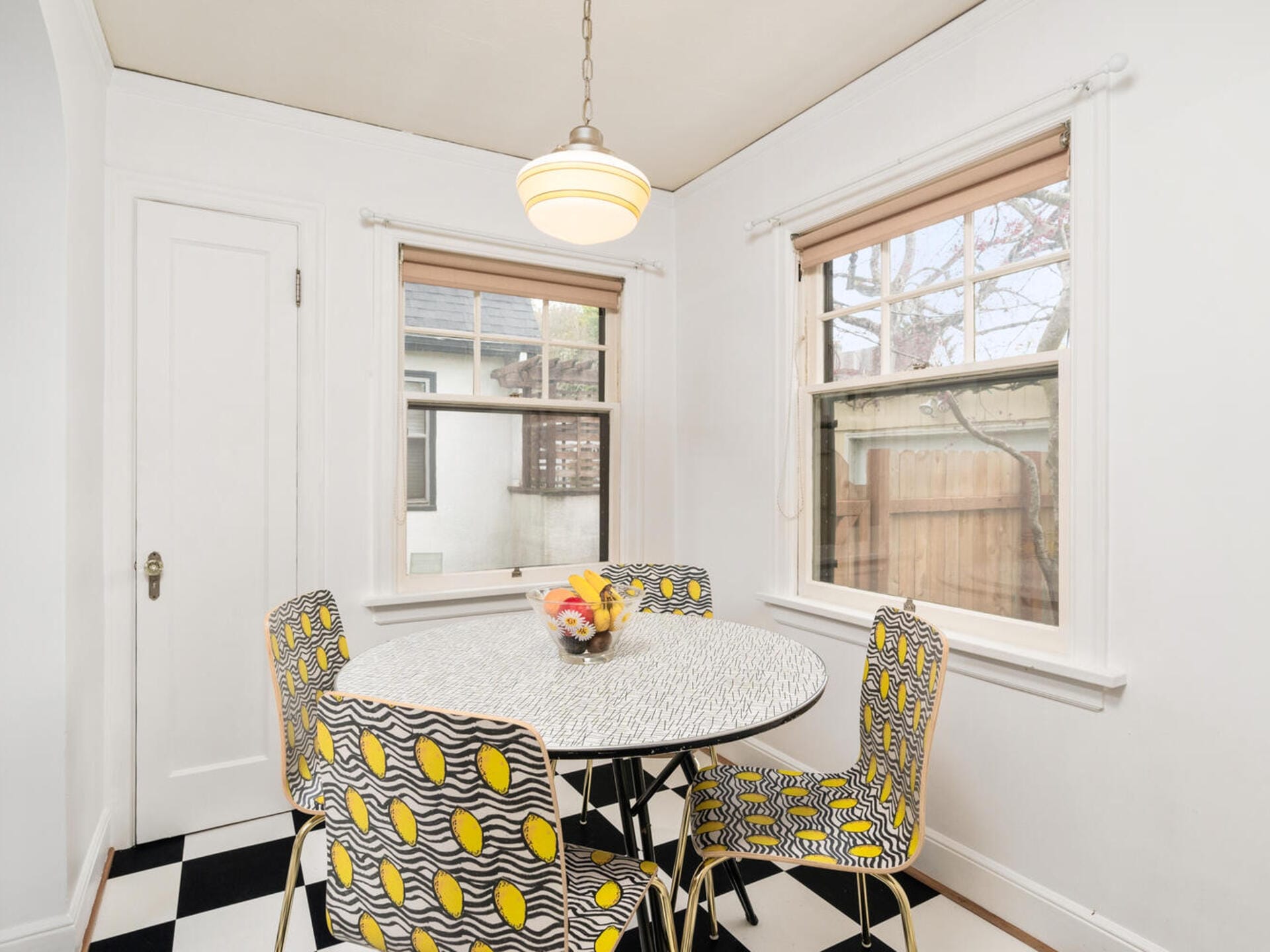 A small dining area with a round table and six chairs. The chairs have black and white geometric patterns with yellow accents. Two windows provide natural light, and the floor features a black and white checkered pattern. A pendant light hangs above.