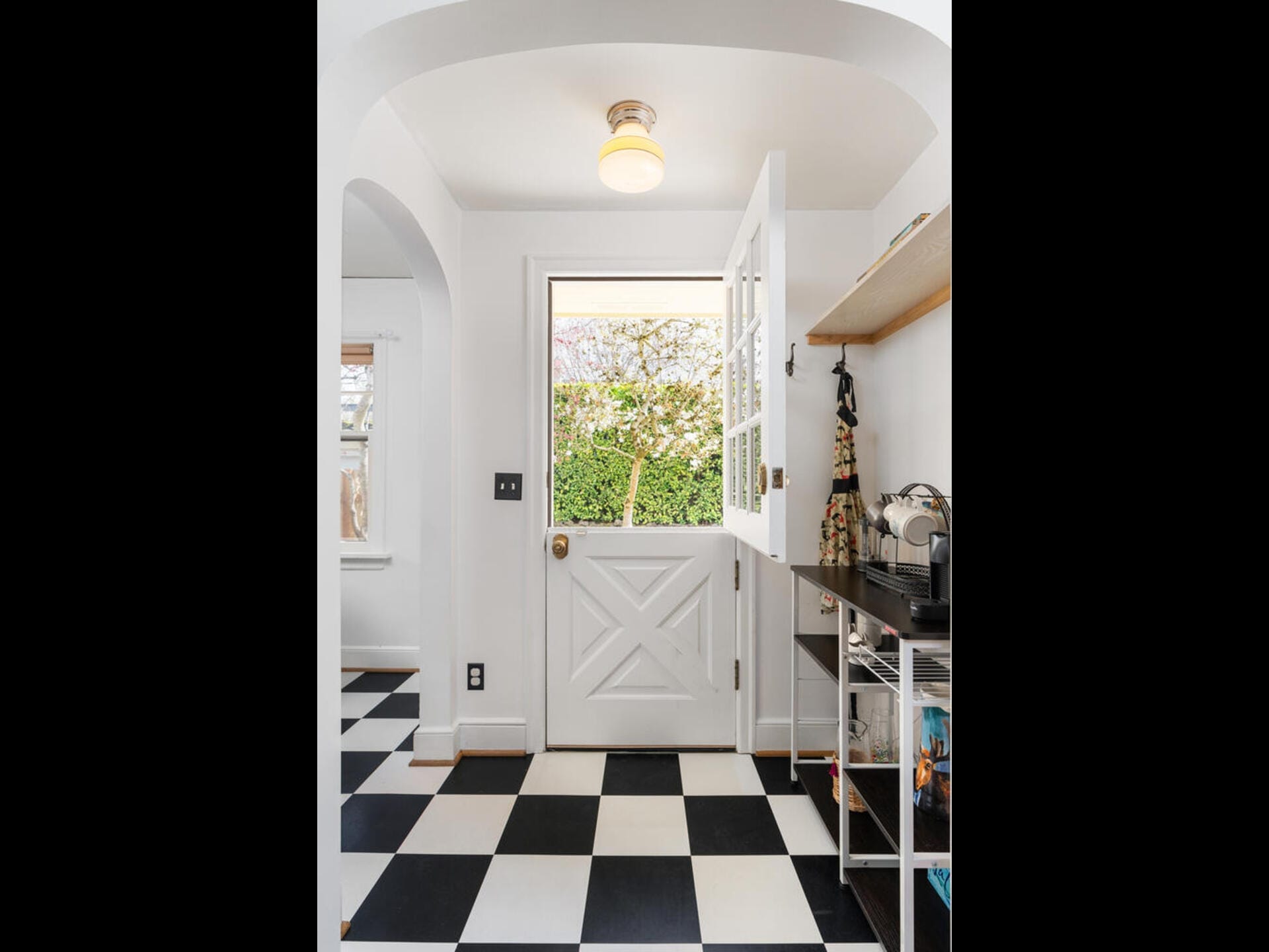 A bright kitchen with a black and white checkered floor. A white Dutch door is open, revealing greenery outside. Inside, there is a countertop with kitchen items and a dress hanging. An overhead light fixture is on the ceiling.