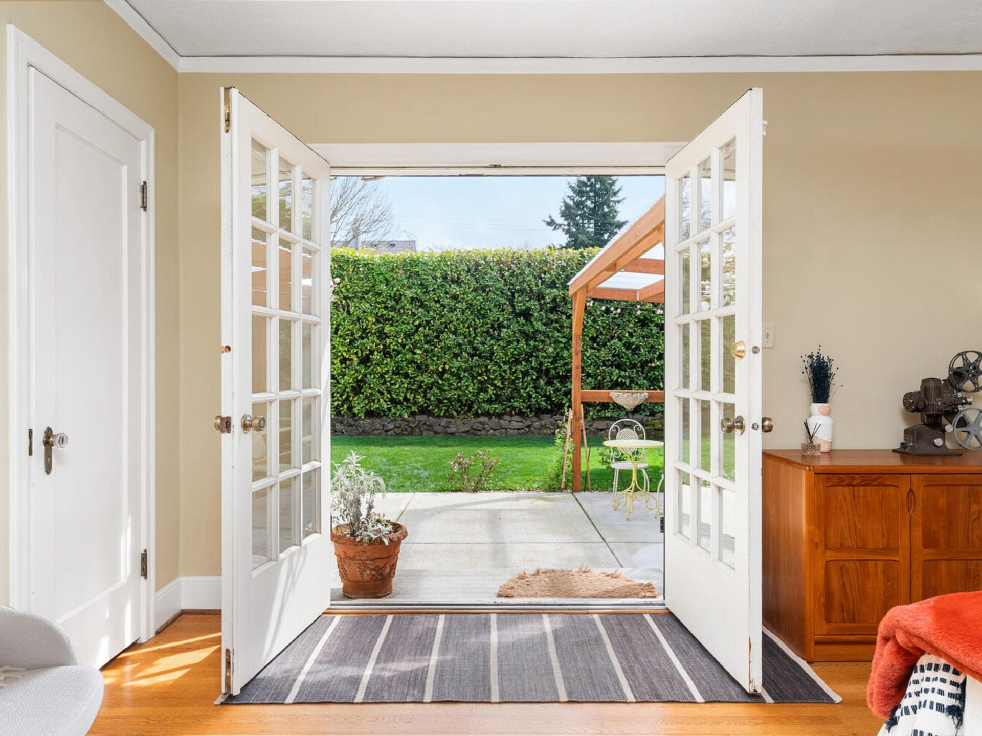 A cozy room with open French doors leading to a patio and lush green garden. A wooden sideboard holds decorative items on the right. A striped rug lies on the wooden floor, and a potted plant is by the door. Natural light fills the space.