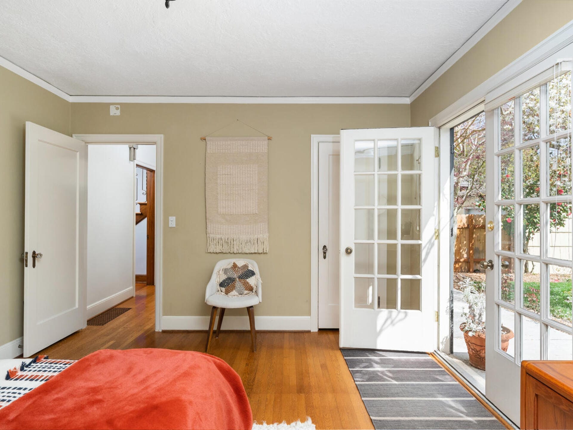 A cozy bedroom with a beige color scheme features a bed with an orange blanket, a white chair with a patterned cushion, and an open door leading to a garden. Sunlight streams through glass doors, illuminating the wooden floor.
